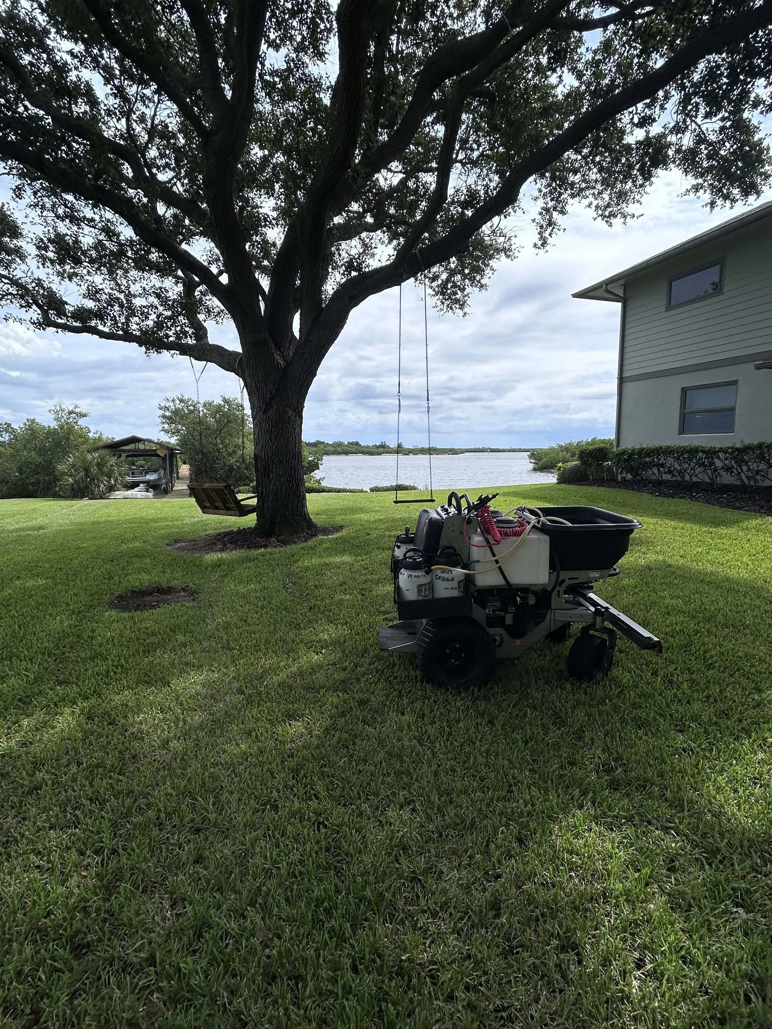 Lawn care equipment unit on grass near a large tree with a swing hanging from it, overlooking a body of water with a house to the right and a carport in the background.