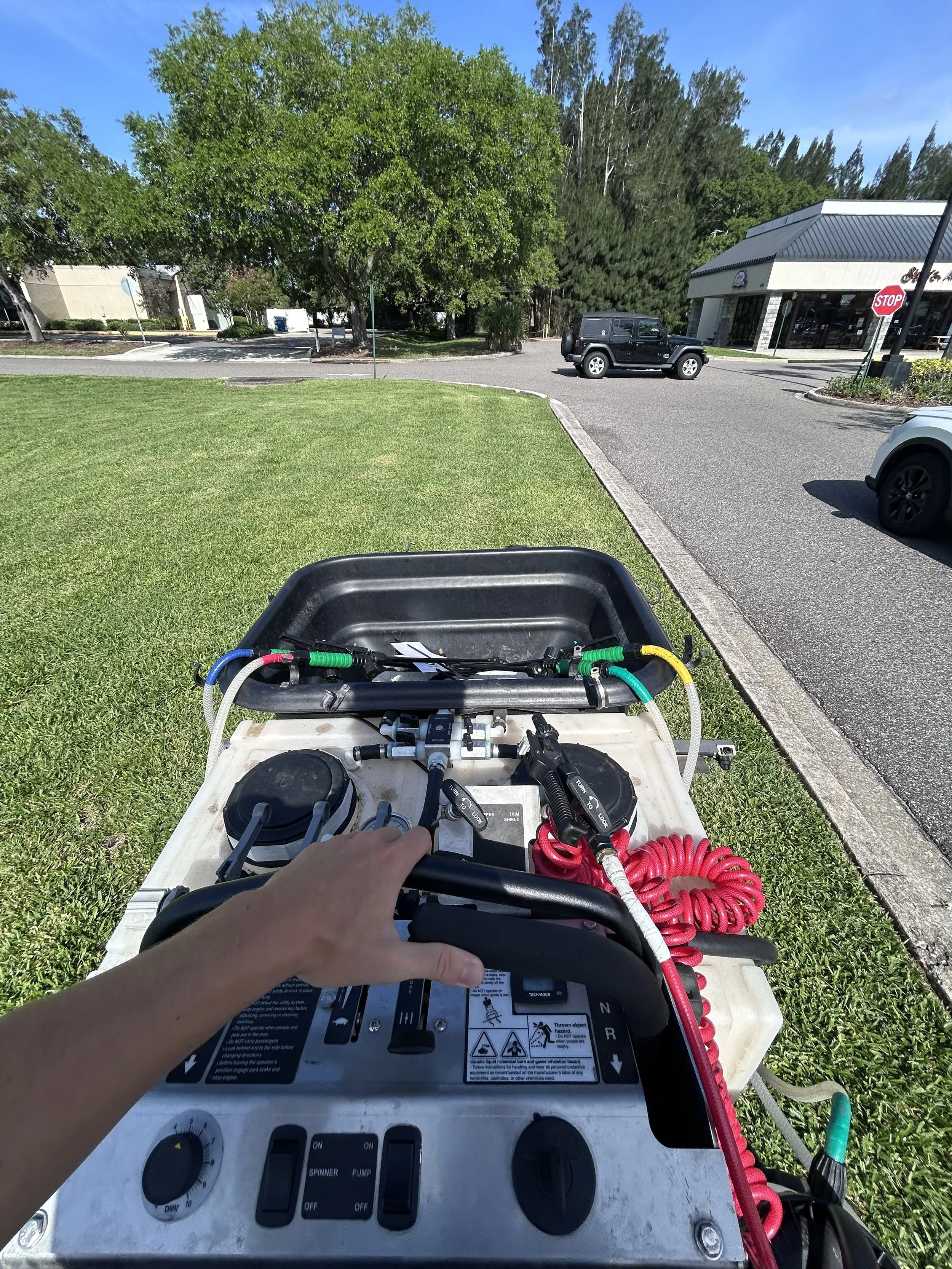 Close-up of a person's hand on the control panel of a portable water or fire suppression pump, with hoses and gauges, outdoors on a grassy area near a parking lot and trees.