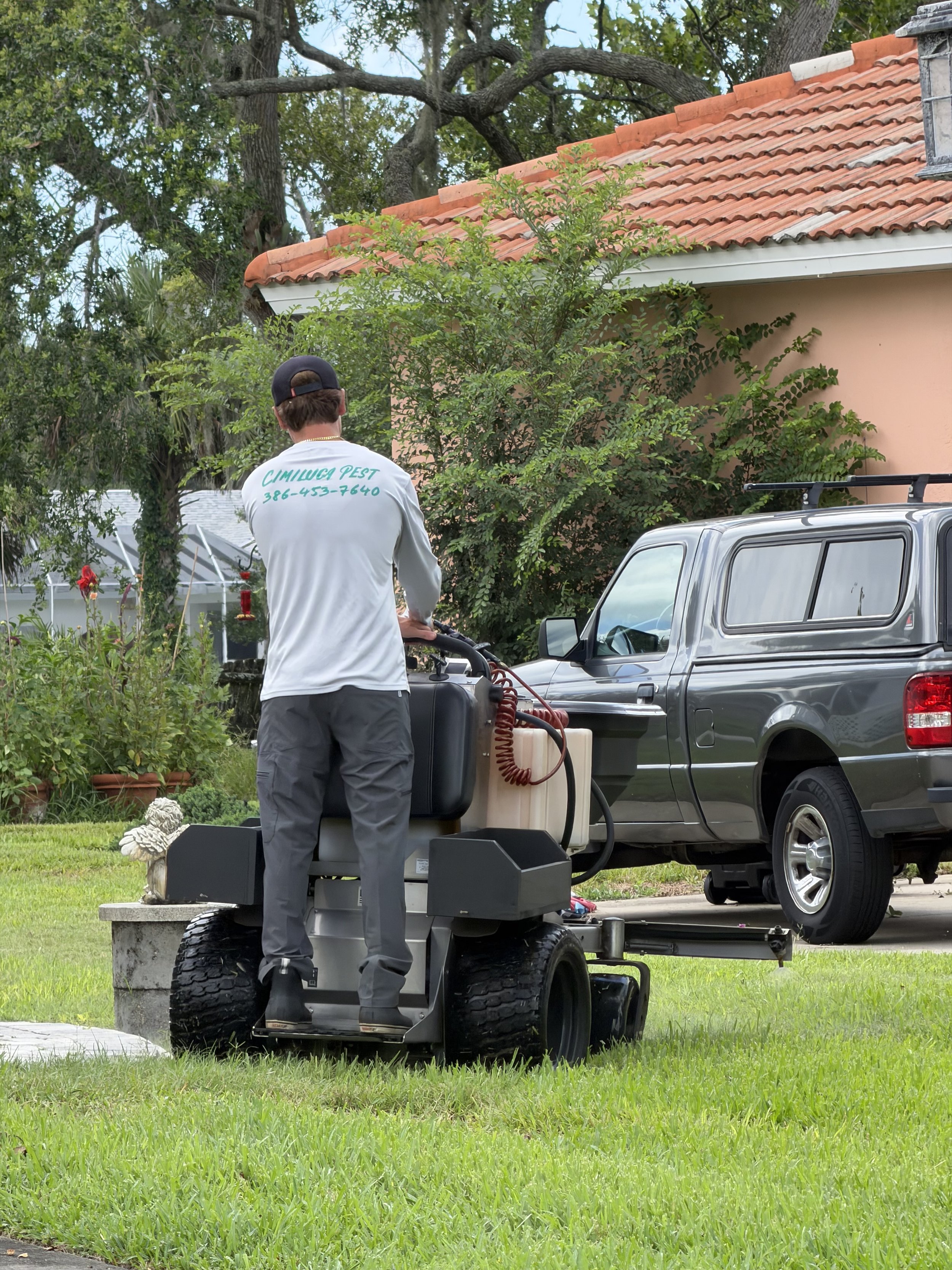 A man in a white shirt with green text and a black cap operating a riding lawn mower on a lush green lawn in front of a house with a red-tiled roof and greenery.