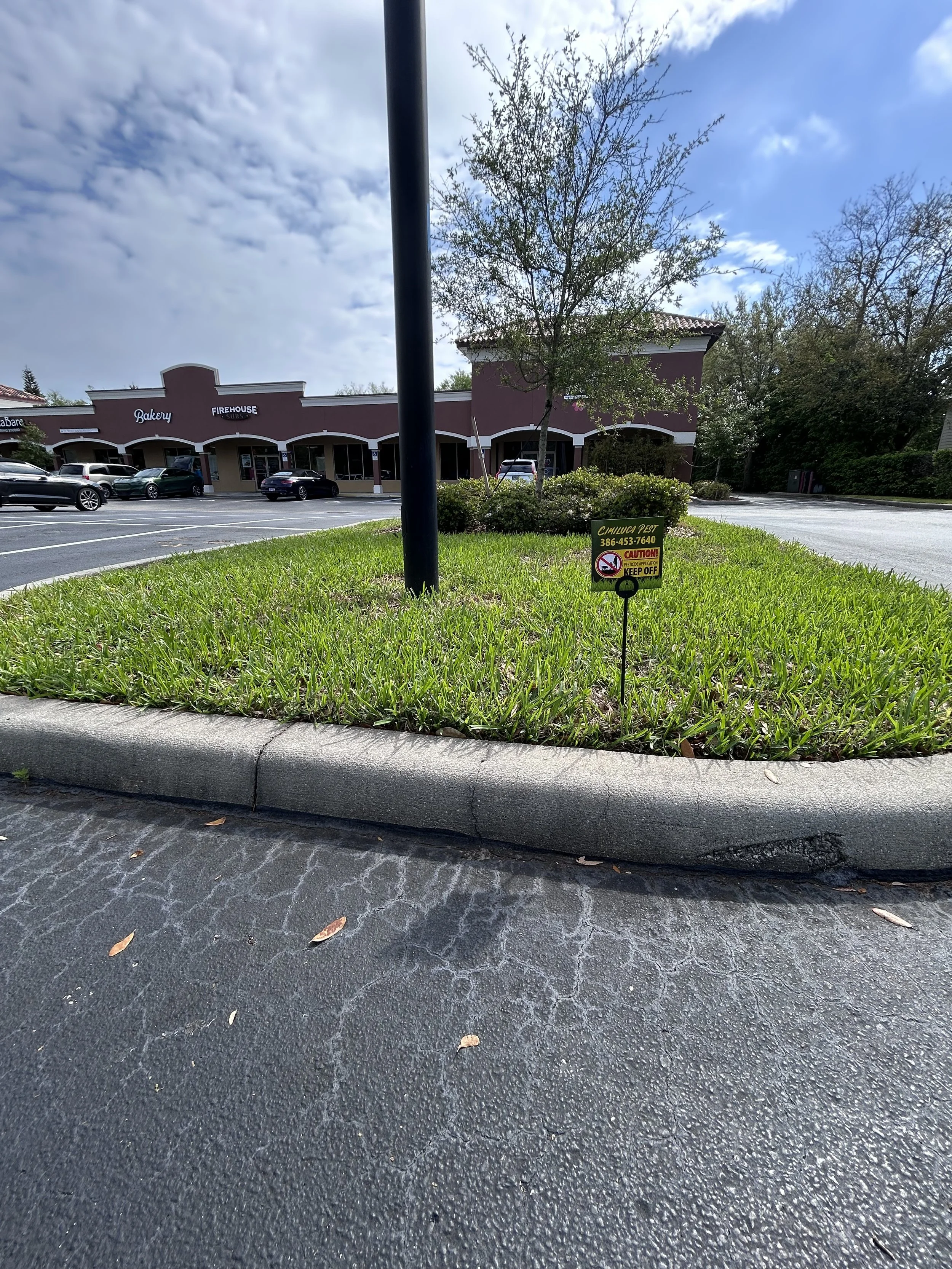 A parking lot with a small grassy island containing a tree, a black pole, and a sign that reads ‘Caution! Keep Off!’ in front of a strip mall with storefronts labeled Bakery and Firehouse.