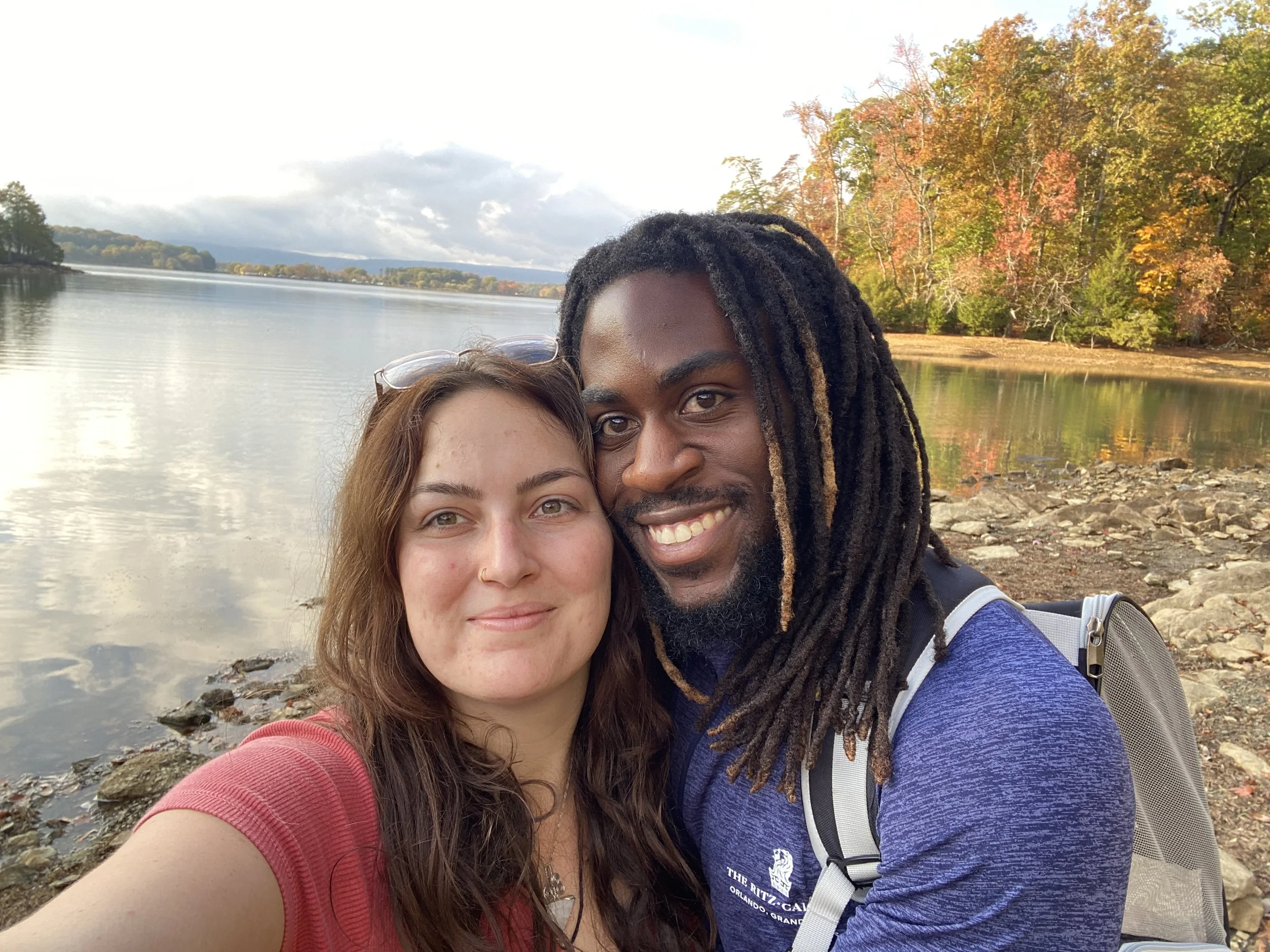 A smiling couple taking a selfie near a calm lake surrounded by trees with fall foliage, with mountains and cloudy sky in the background.