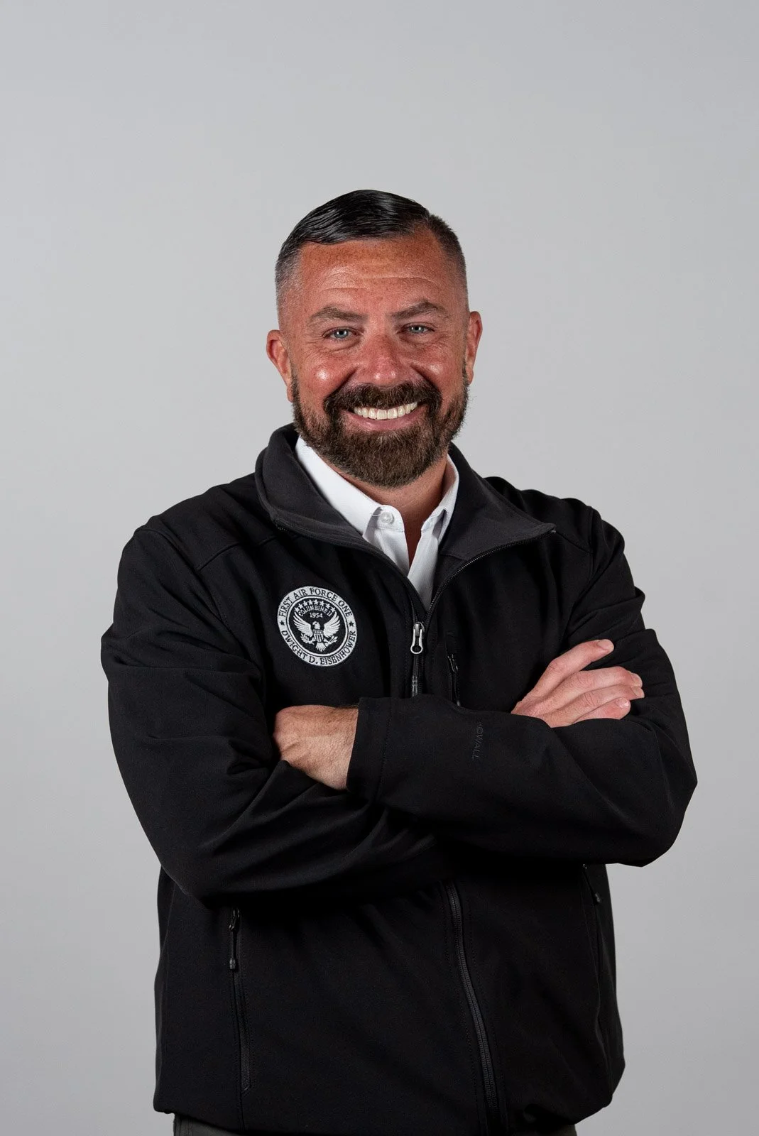 A smiling man with a beard and short hair wearing a black jacket with a patch and a white shirt underneath, standing with his arms crossed against a plain gray background.