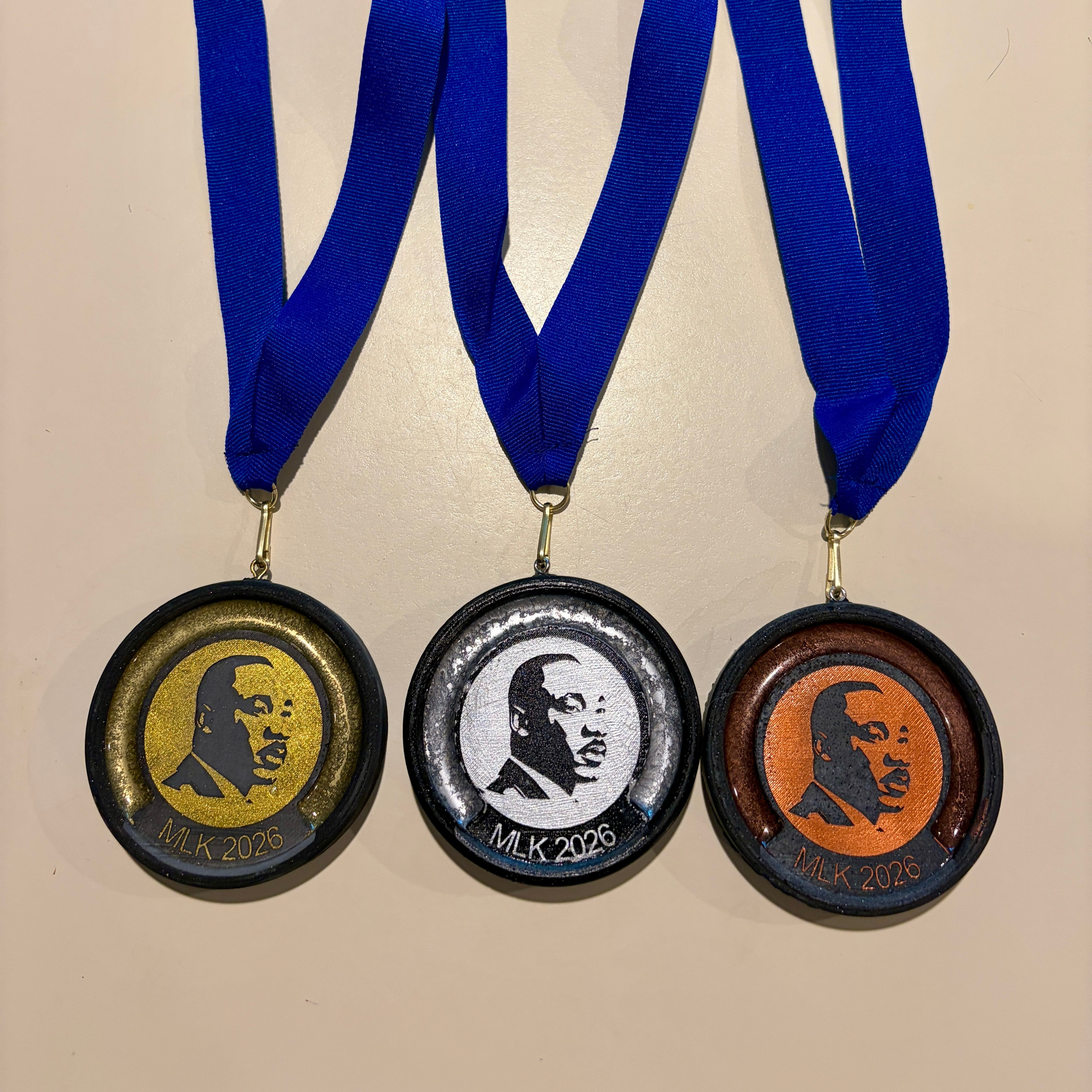 Three medals hanging from blue ribbons. The medals feature a silhouette of Martin Luther King, Jr. with the text "MLK 2026" at the bottom, in three different colors: gold, silver, and bronze.