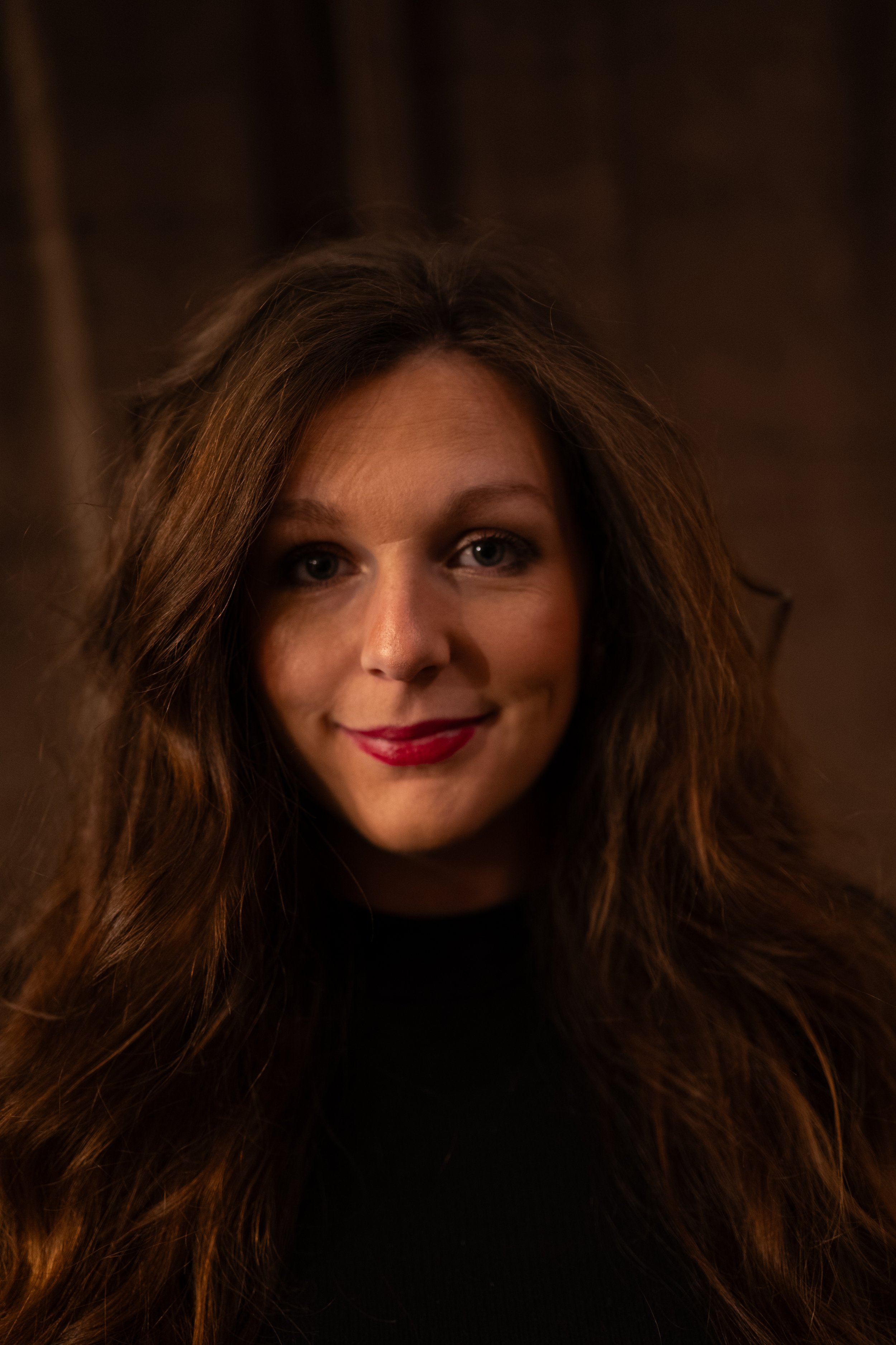 A woman with long wavy brown hair, bright eyes, and red lipstick, smiling warmly, wearing a black top, with a blurred dark background.