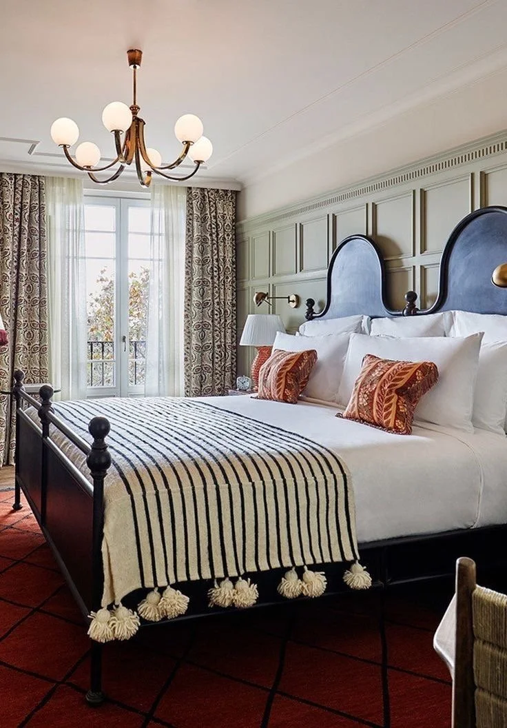 A neatly made bed with white linens, striped black and white throw blanket, and decorative orange pillows in a bedroom with a chandelier, wall lamps, and large windows with patterned curtains.