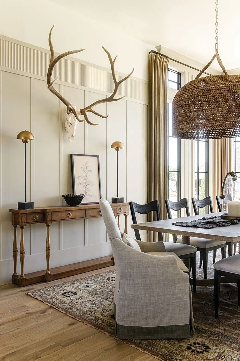 A dining room with a large antler chandelier hanging above a wooden dining table surrounded by black and beige chairs. The room has tall windows with beige curtains, a wooden sideboard with brass lamps, a framed botanical print, a decorative bowl, and a rug with traditional patterns on the wooden floor.