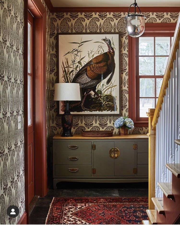 A hallway corner featuring a green sideboard with brass handles and hardware, a white table lamp, a wooden tray, and a vase with blue hydrangeas. Above, a large framed painting of a pheasant hangs on front of vintage-style paisley wallpaper. To the right, there is a wooden staircase with white balusters and a yellow handrail, and a window with a view of trees outside. A glass pendant light hangs from the ceiling.