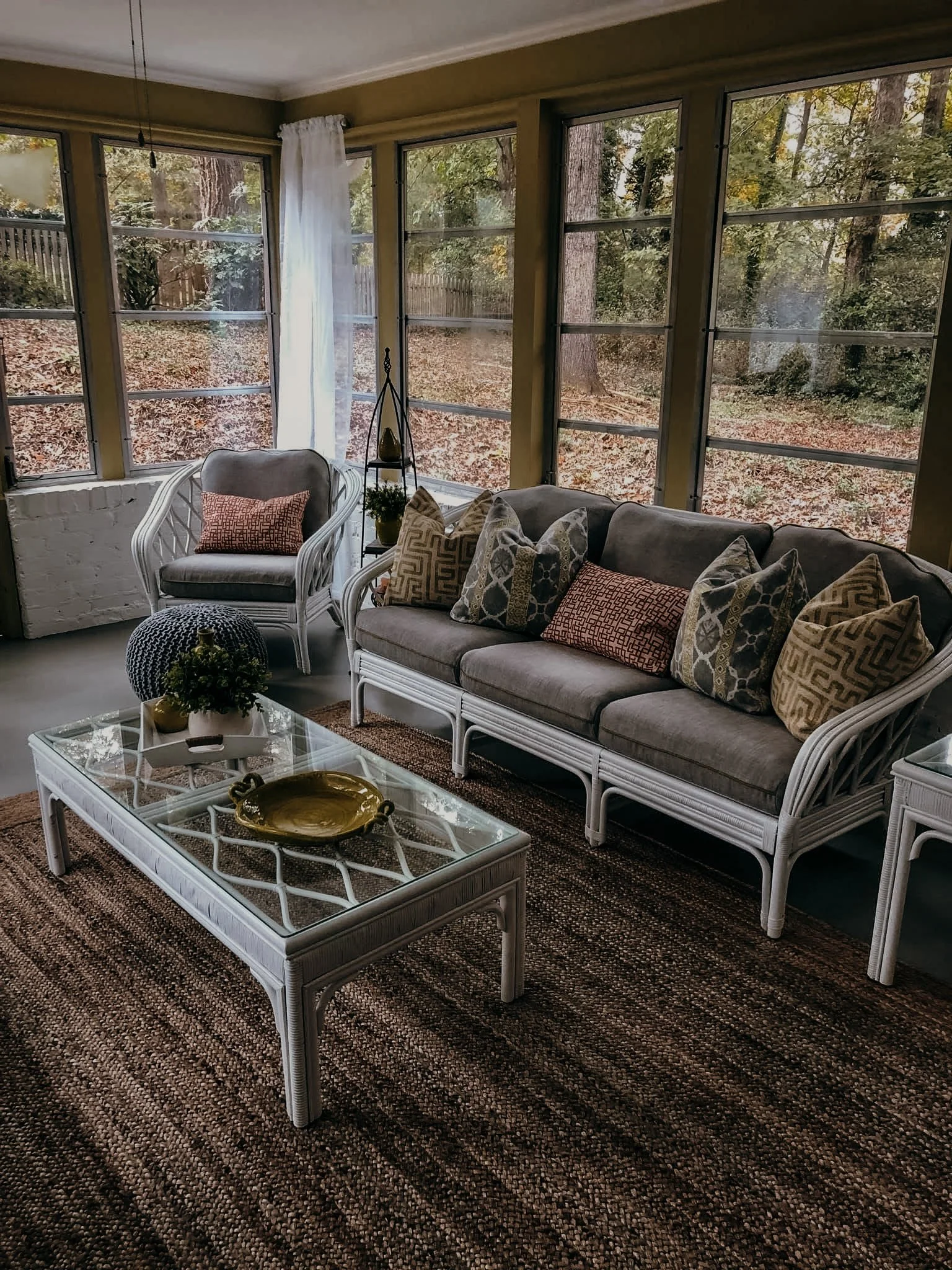 Cozy living room with large windows, white wicker furniture, a glass coffee table, patterned throw pillows, potted plants, and an earthy-toned rug, overlooking a wooded yard.