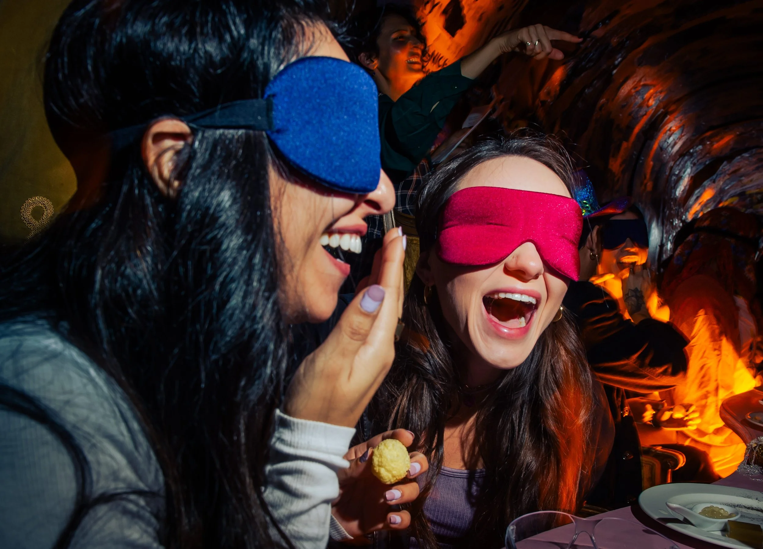 Two guests laughing and wearing colorful blindfolds laugh during an interactive participatory experience.
