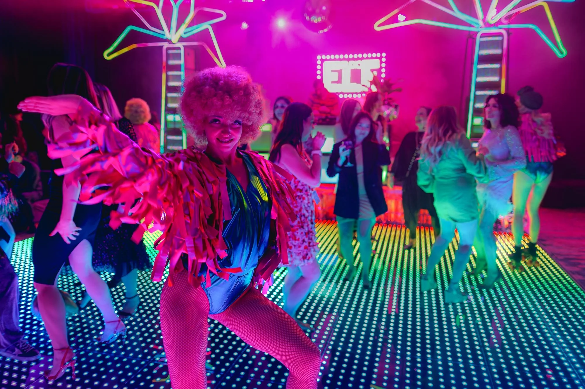 Costumed performer leading guests across an illuminated dance floor inside a colorful immersive experience.