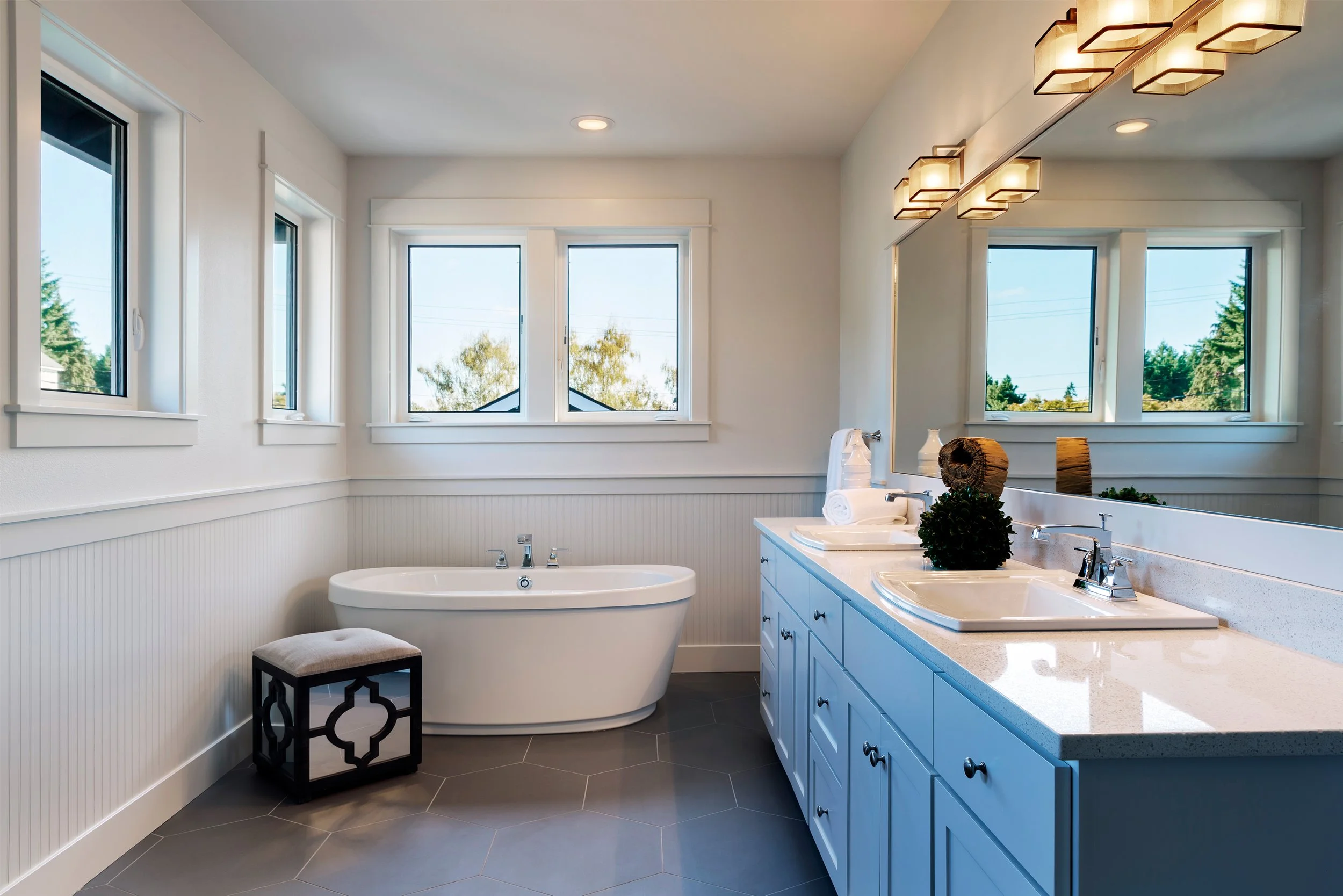 Bright bathroom with a white bathtub, a blue vanity with white countertop, a large mirror, three windows showing trees outside, and decorative items on the counter.