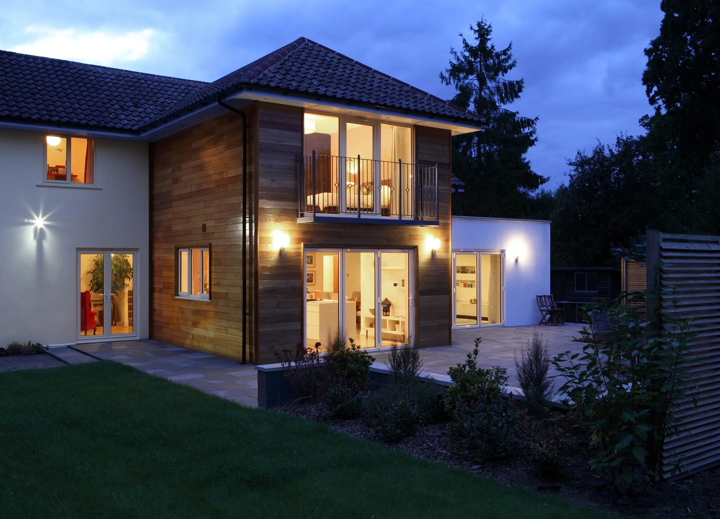 A modern two-story house illuminated at dusk, featuring a wooden exterior and large glass doors and windows, with a balcony on the second floor and a landscaped yard.