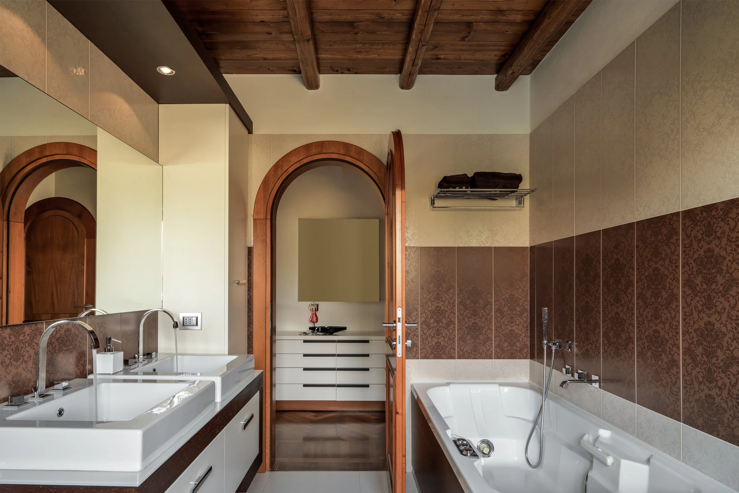 Bathroom with double sink vanity, large mirror, bathtub, wooden ceiling, arched doorway, and towel rack.