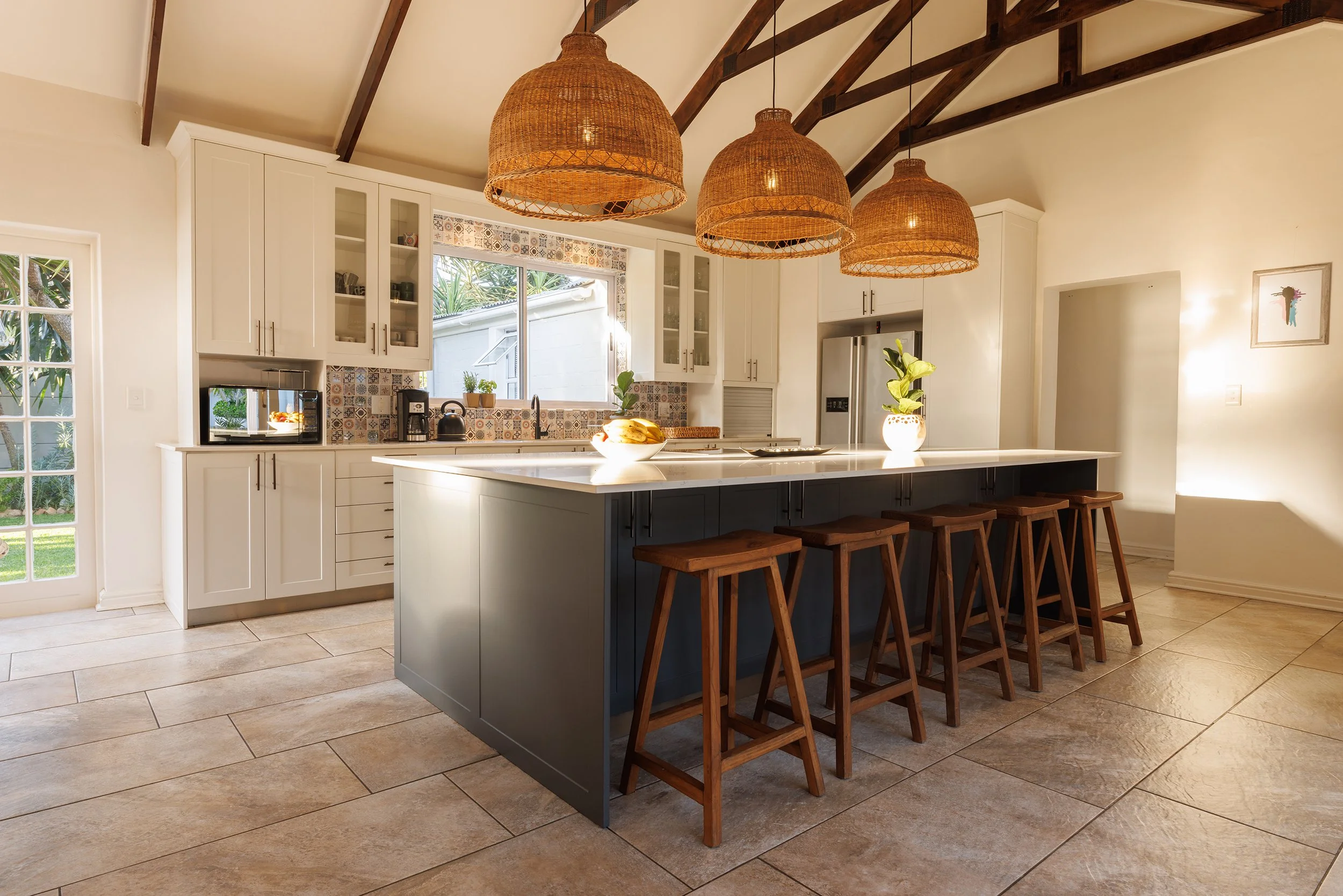 Modern kitchen with white cabinets, a large island with a dark base, wooden bar stools, woven pendant lights, tiled backsplash, and a window overlooking a garden.