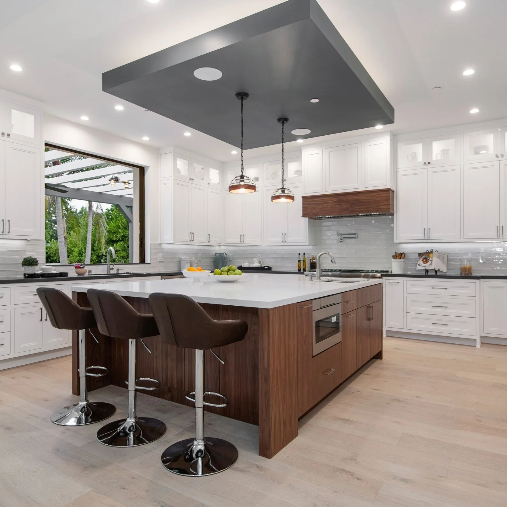 Modern kitchen with white cabinets a large central island with a wooden base and white countertop, three brown bar stools, and pendant lights hanging above. There is a window with a view of an outdoor patio and green trees.