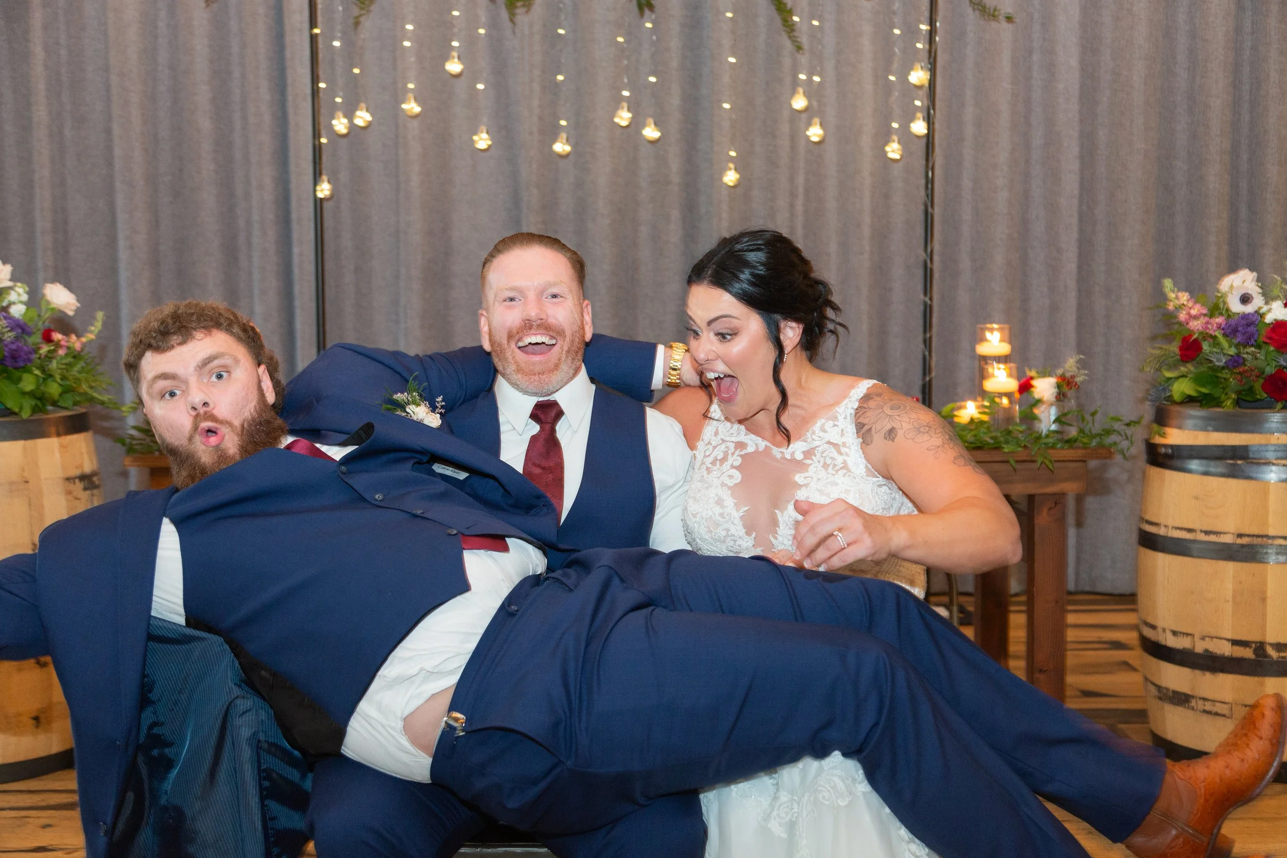 Three people in formal wedding attire having fun and posing playfully at a wedding reception, with floral decorations and candles in the background.