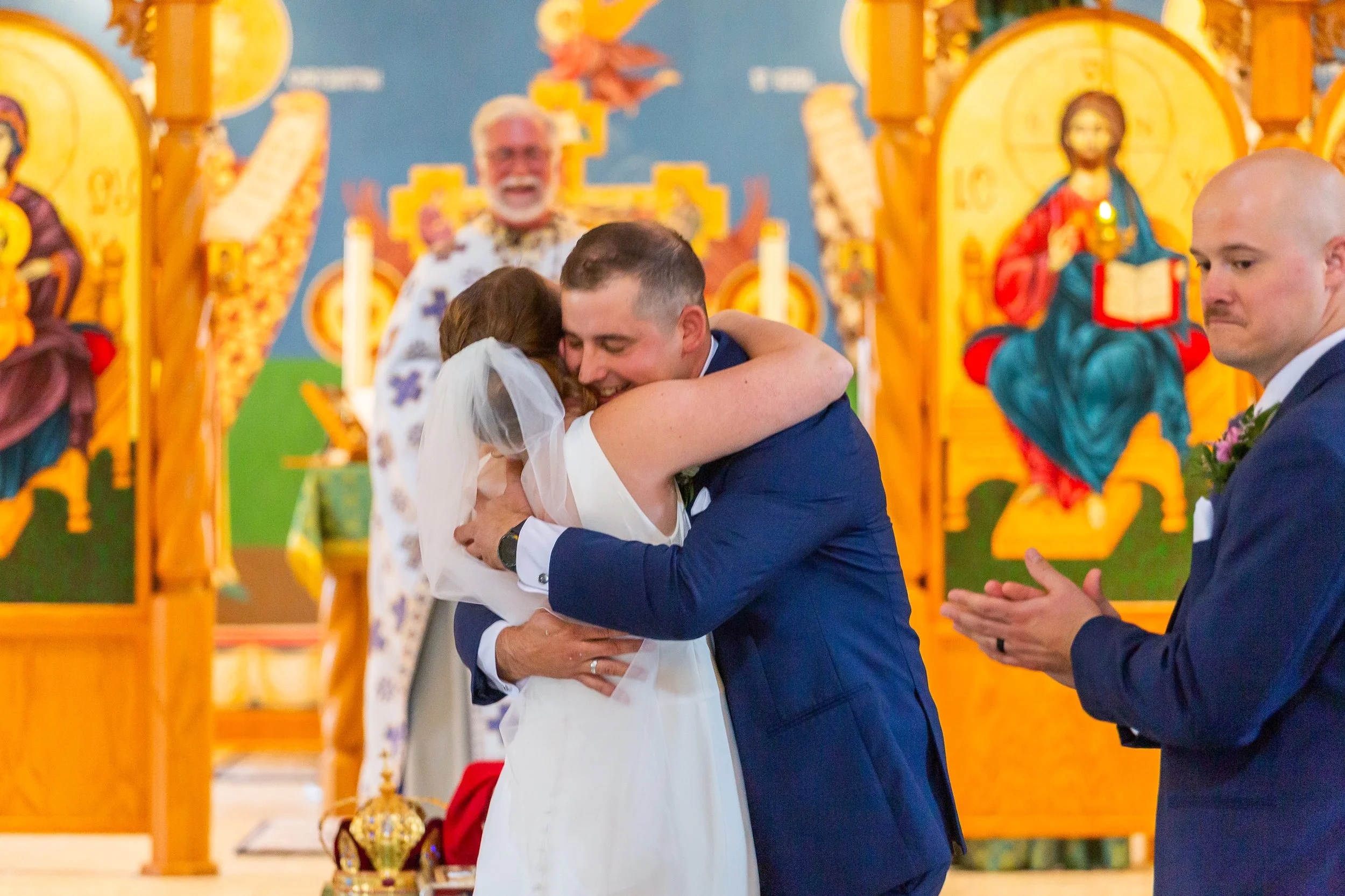 A bride and groom hugging during a wedding ceremony in a church with religious icons in the background. A officiant and a man clapping are also visible.