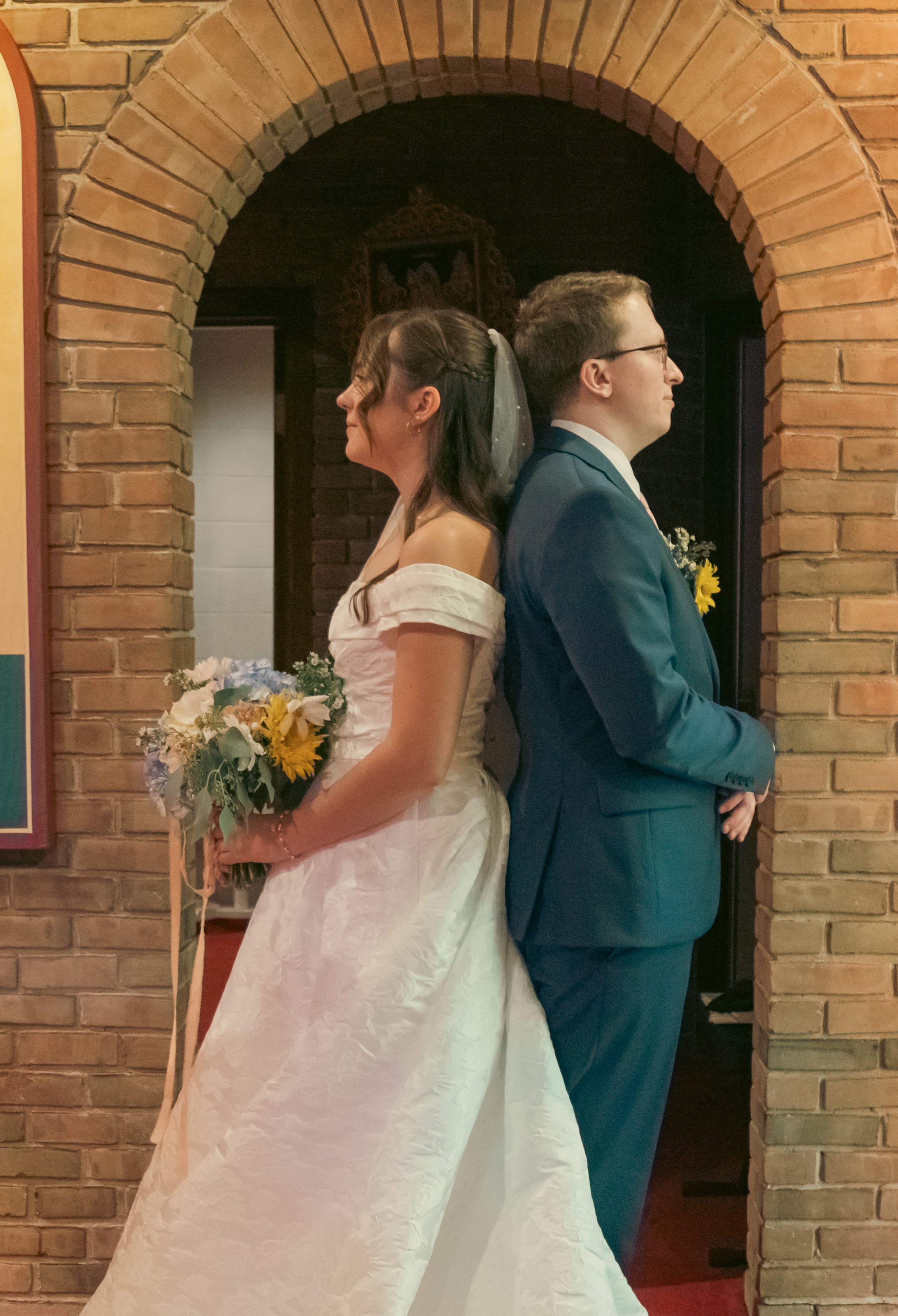A bride and groom stand back to back, leaning against each other with their backs touching, in a brick archway during their wedding ceremony.