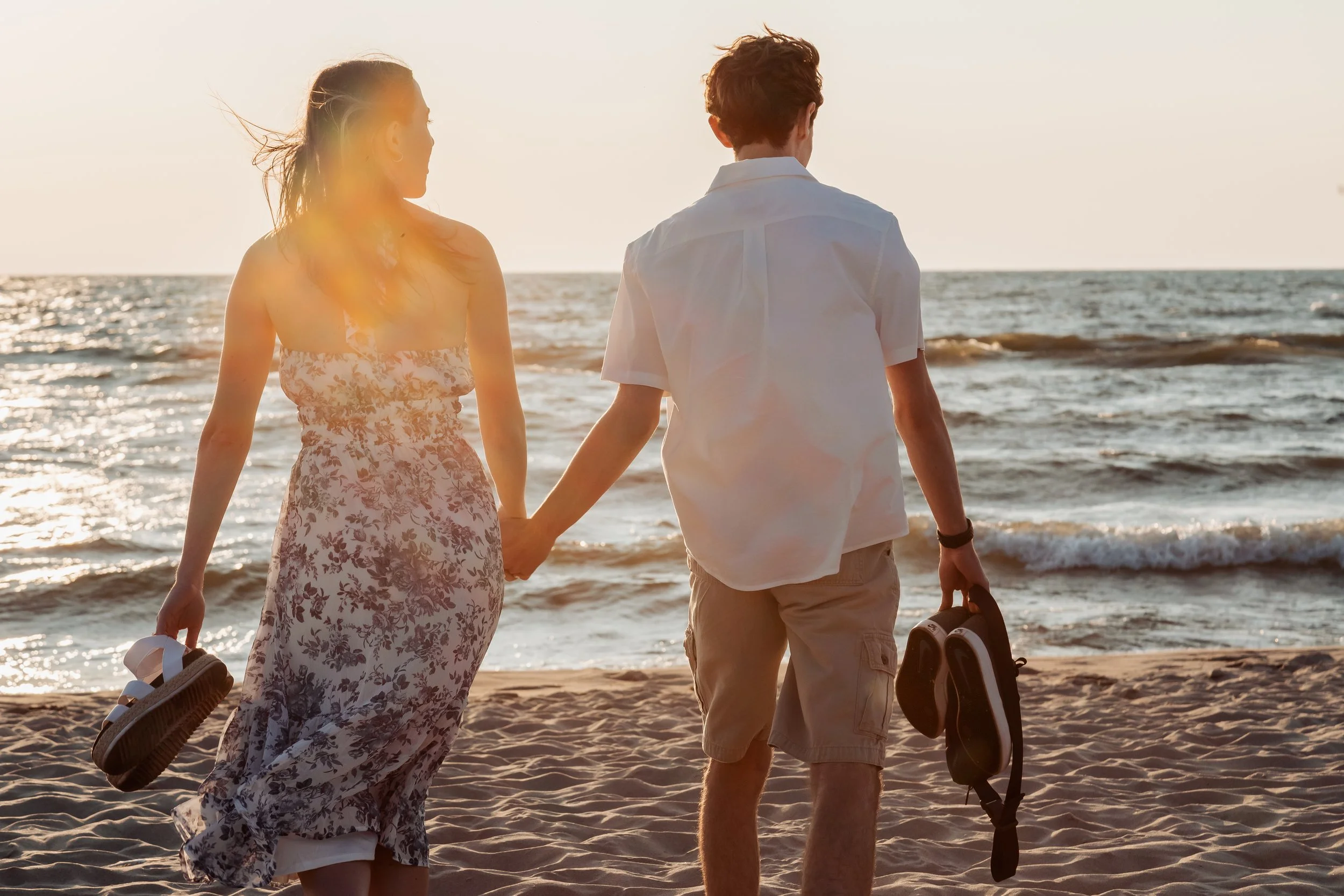 A young couple holding hands and walking on a beach at sunset, each carrying a sandal or a shoe, with the ocean and waves in the background.