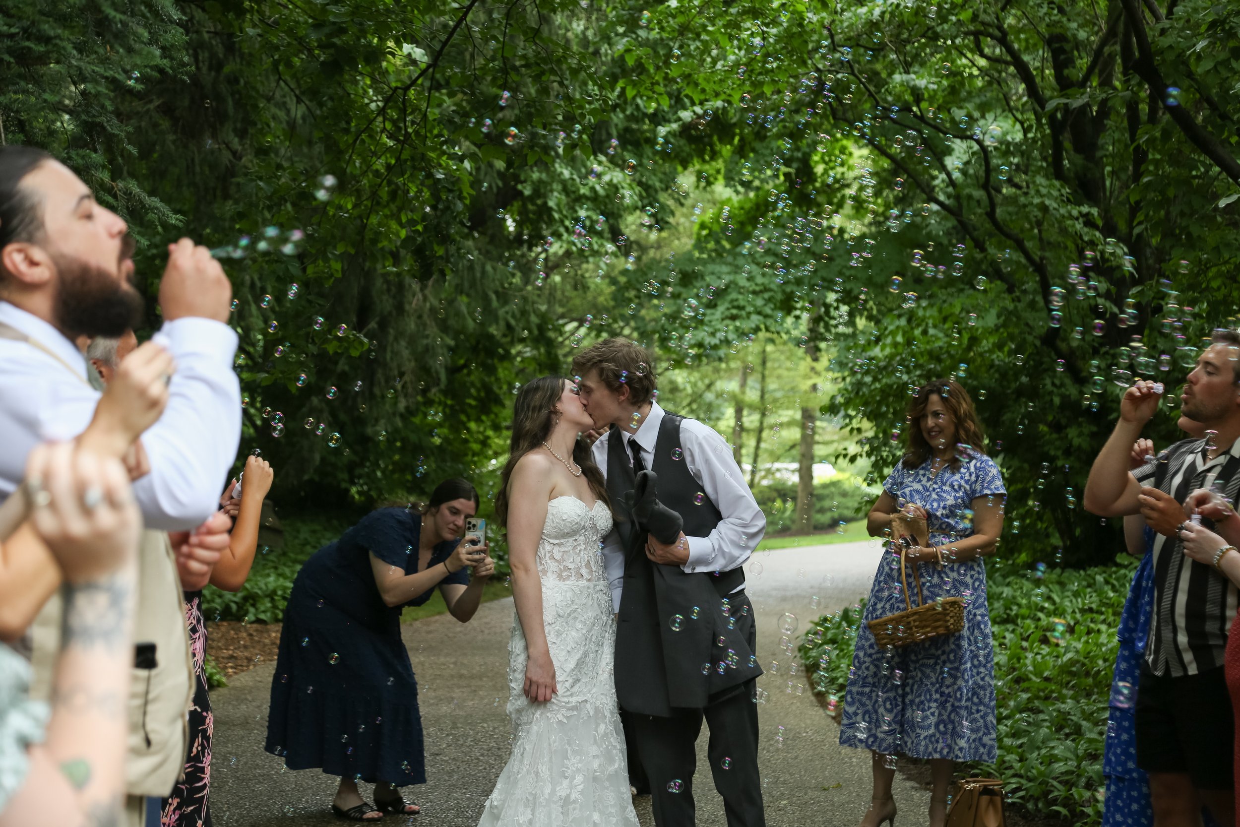 A wedding couple sharing a kiss surrounded by friends and family in a wooded outdoor setting, with bubbles floating in the air.