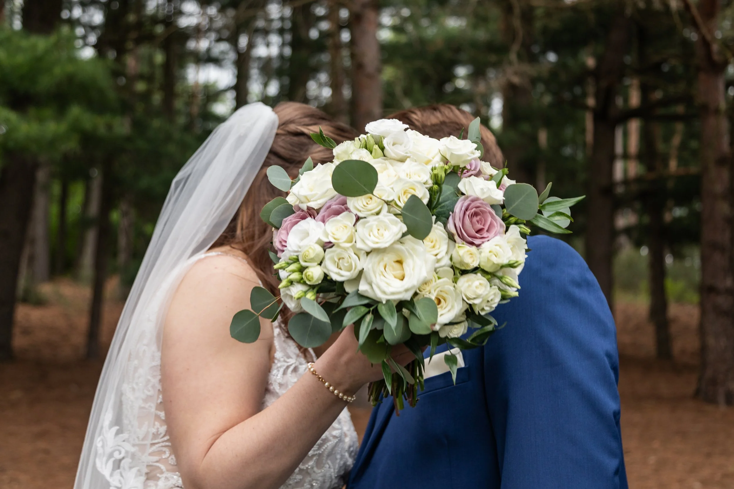 Bride holding a bouquet of white and pink roses, partially covering her face, with groom in a blue suit, in a forest setting.