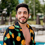 A young man with a beard smiling outdoors, wearing a black shirt with pineapple prints.
