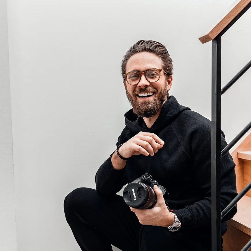 A smiling man with glasses, a beard, and brown hair, sitting on stairs with a camera in his hand, wearing a black hoodie.