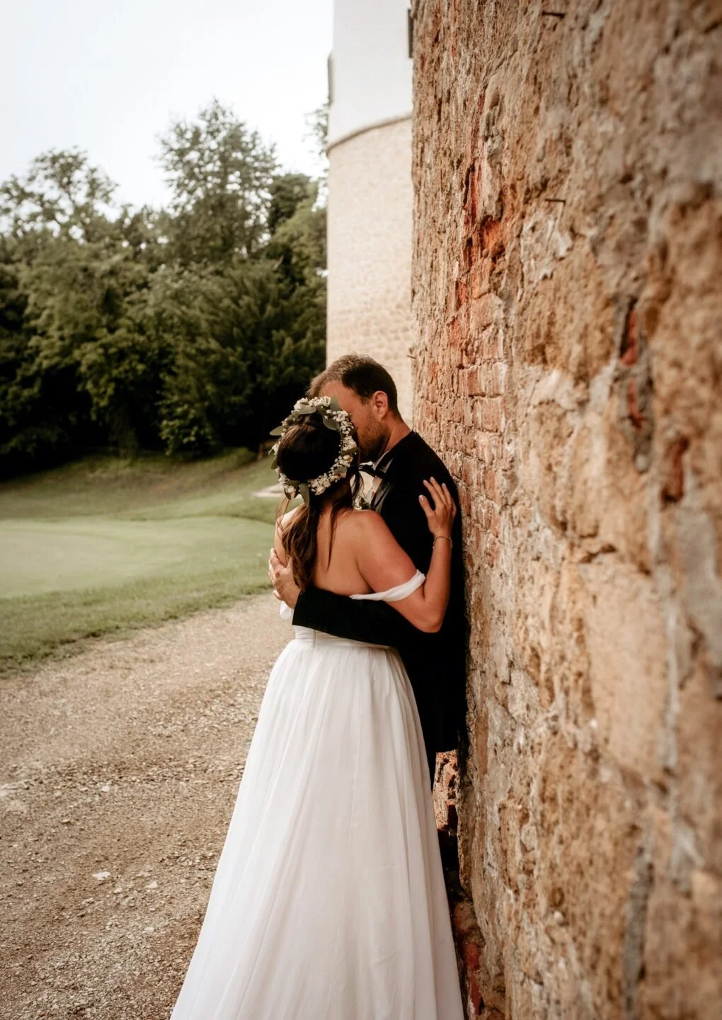 A bride and groom kissing against a brick wall outdoors with greenery in the background.