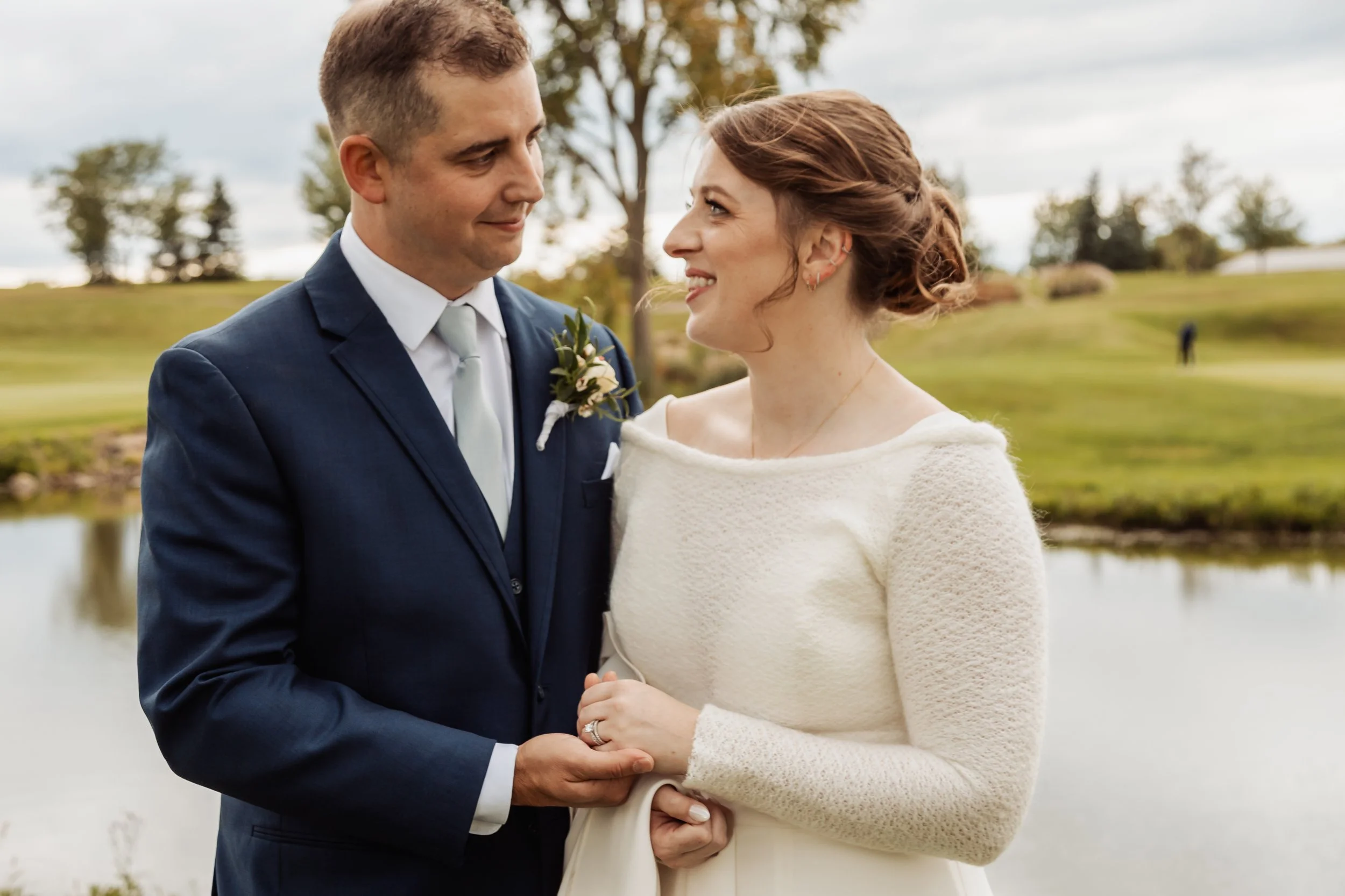 A bride and groom standing outdoors by a pond, gazing at each other and holding hands.