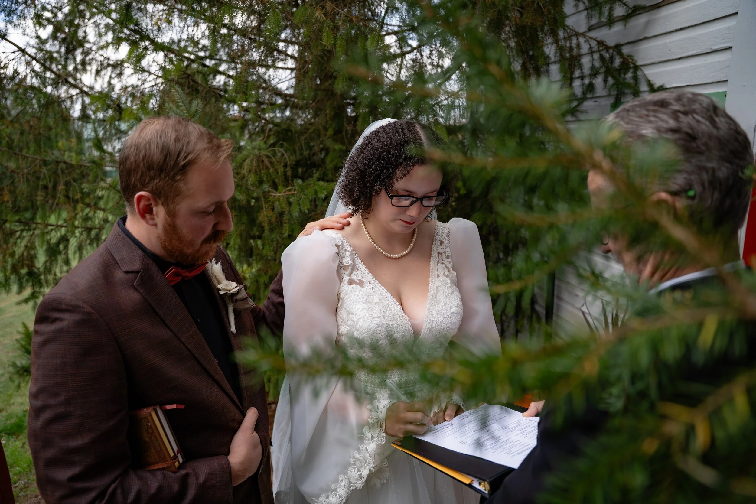 A bride and groom standing outdoors during their wedding ceremony, with a officiant. They are signing a document, with greenery and a white building in the background.