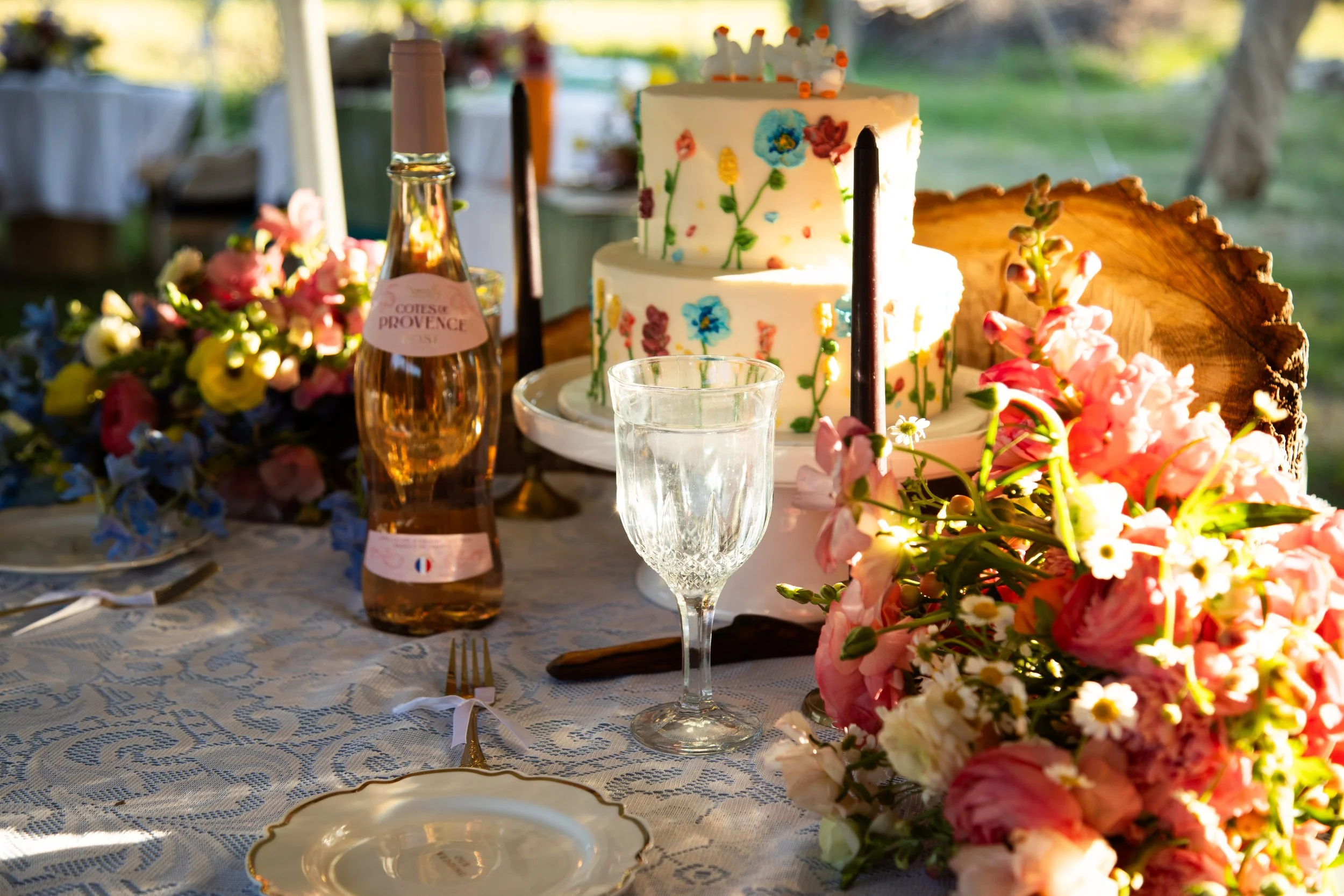 A decorated wedding table with a two-tiered cake with floral designs, a clear crystal glass, pink and white flower arrangements, a bottle of rosé wine, and a plate with a gold rim, set outdoors with green grass and trees in the background.