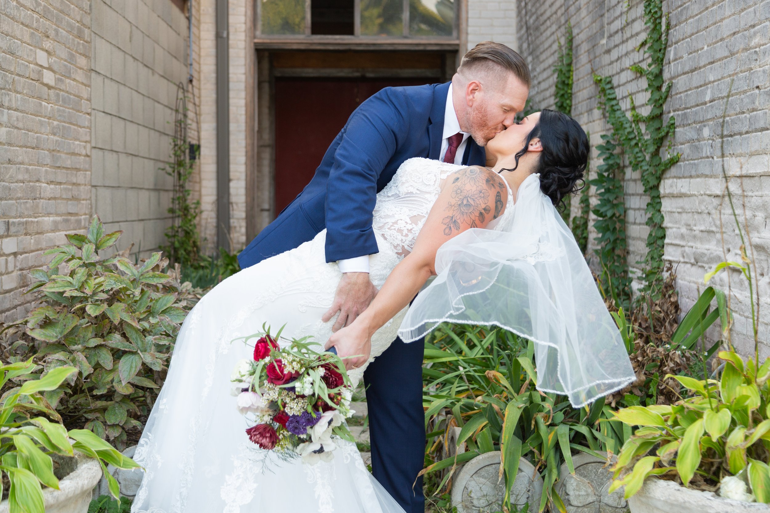A bride and groom share a kiss outdoors, with the groom dipping the bride backward. The bride wears a white wedding dress and veil, holding a colorful bouquet, and has tattoos on her shoulder. The groom wears a blue suit, white shirt, and maroon tie. They are surrounded by plants and brick walls.