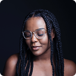 Close-up portrait of a woman with glasses and braided hair, looking downward against a dark background.