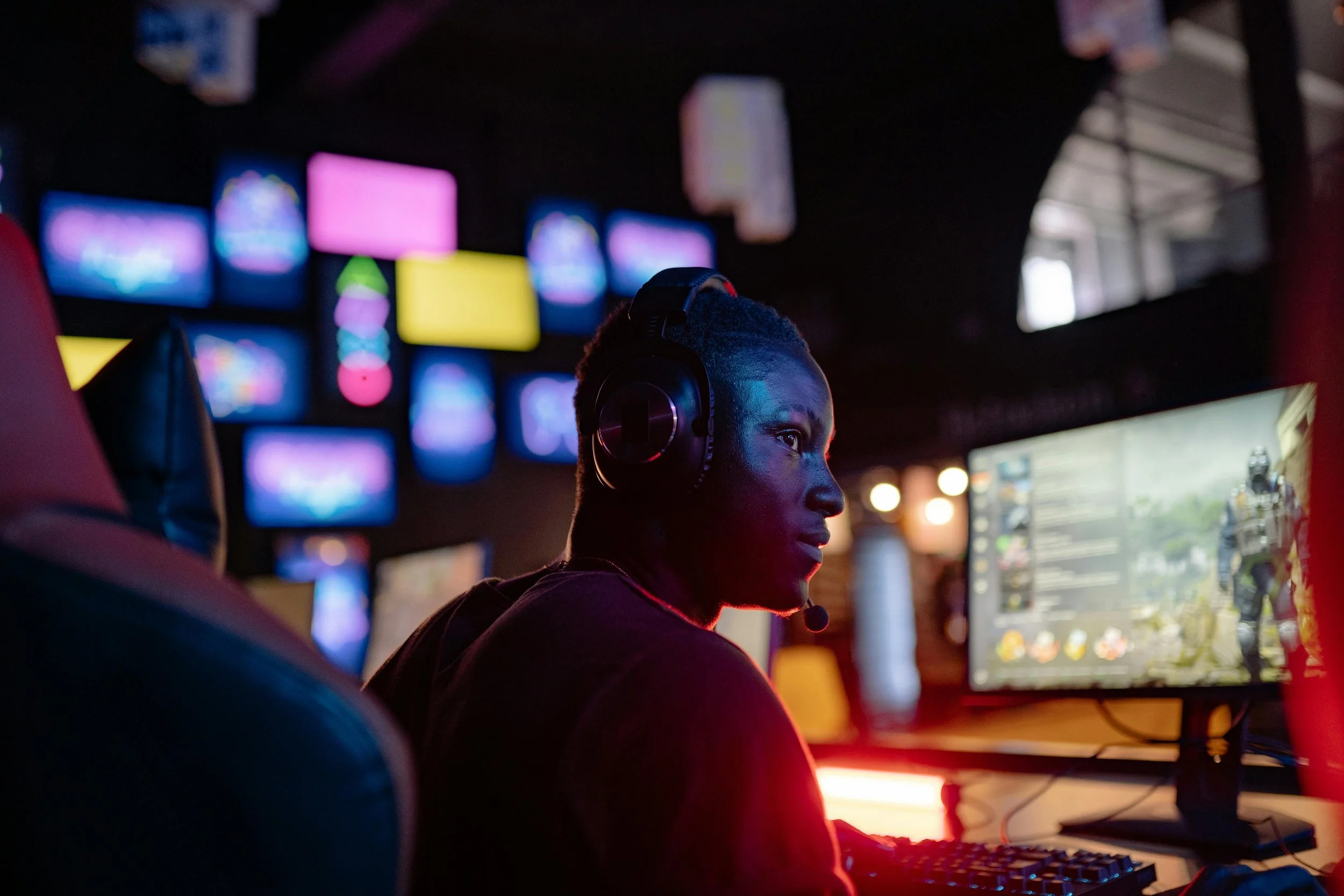 A young man wearing a headset with a microphone is sitting at a computer in a gaming or eSports setting, illuminated by multiple large monitors displaying vibrant colors and graphics.