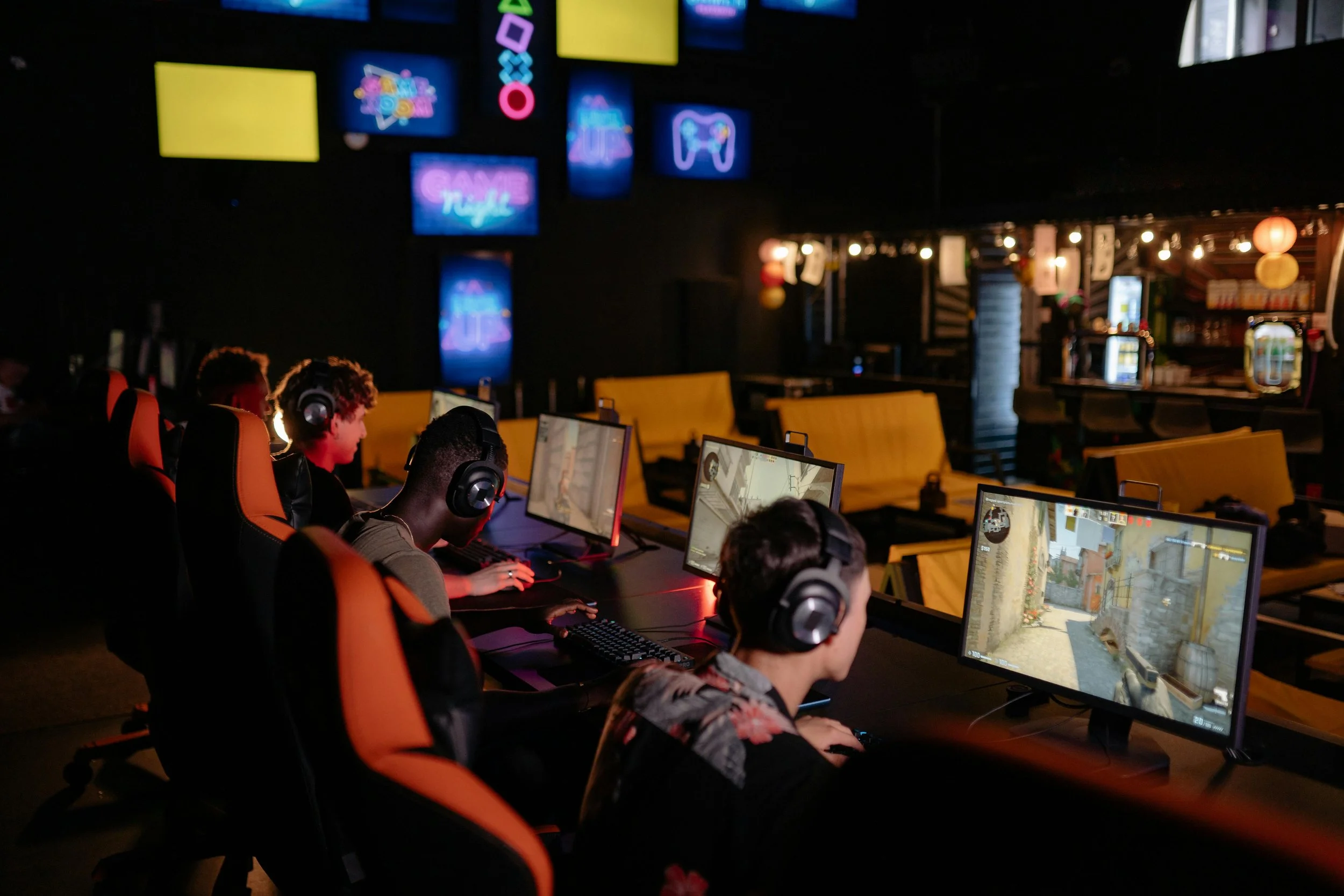 Young people playing video games on computers in a dark gaming cafe or arcade, with neon signs and monitors showing game graphics in the background.