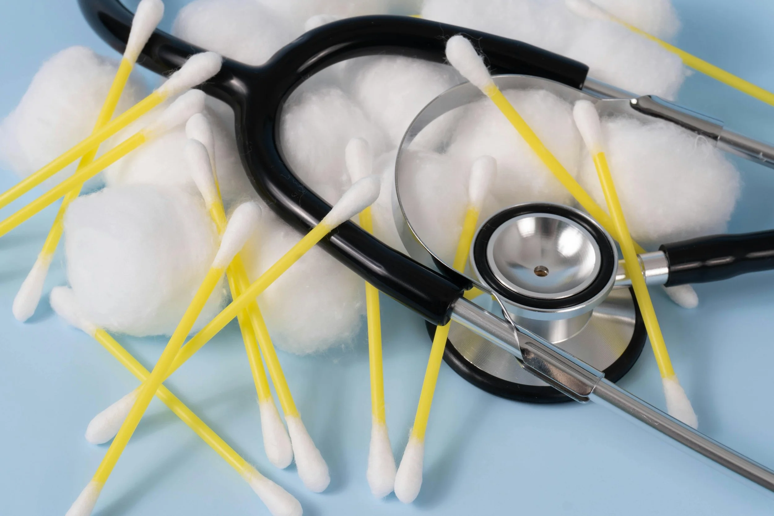 Medical stethoscope placed on top of yellow cotton swabs and cotton balls on a light blue background.