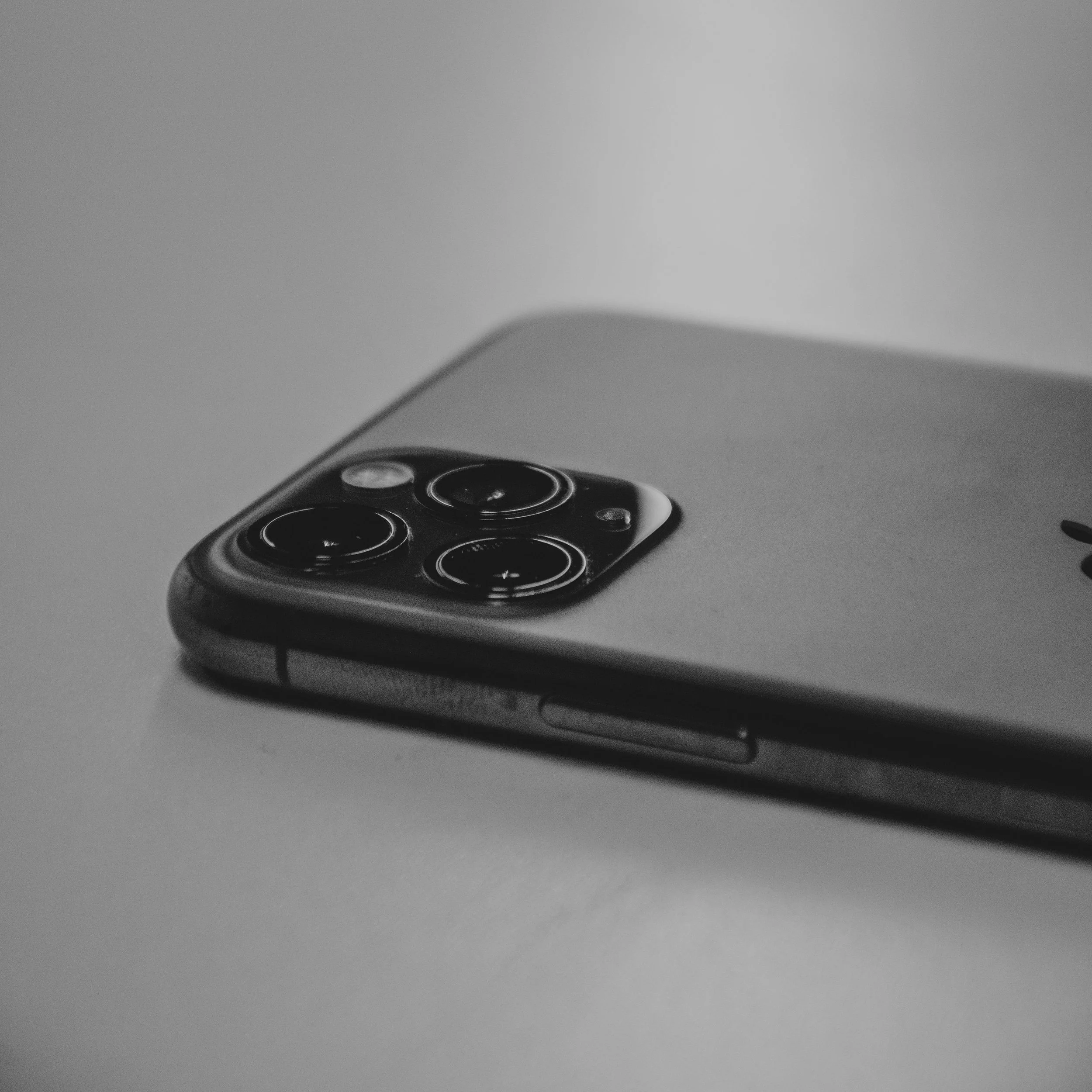 Close-up of a modern smartphone with a camera lens visible, placed on a smooth surface, with part of the screen and rounded edges visible, in grayscale.