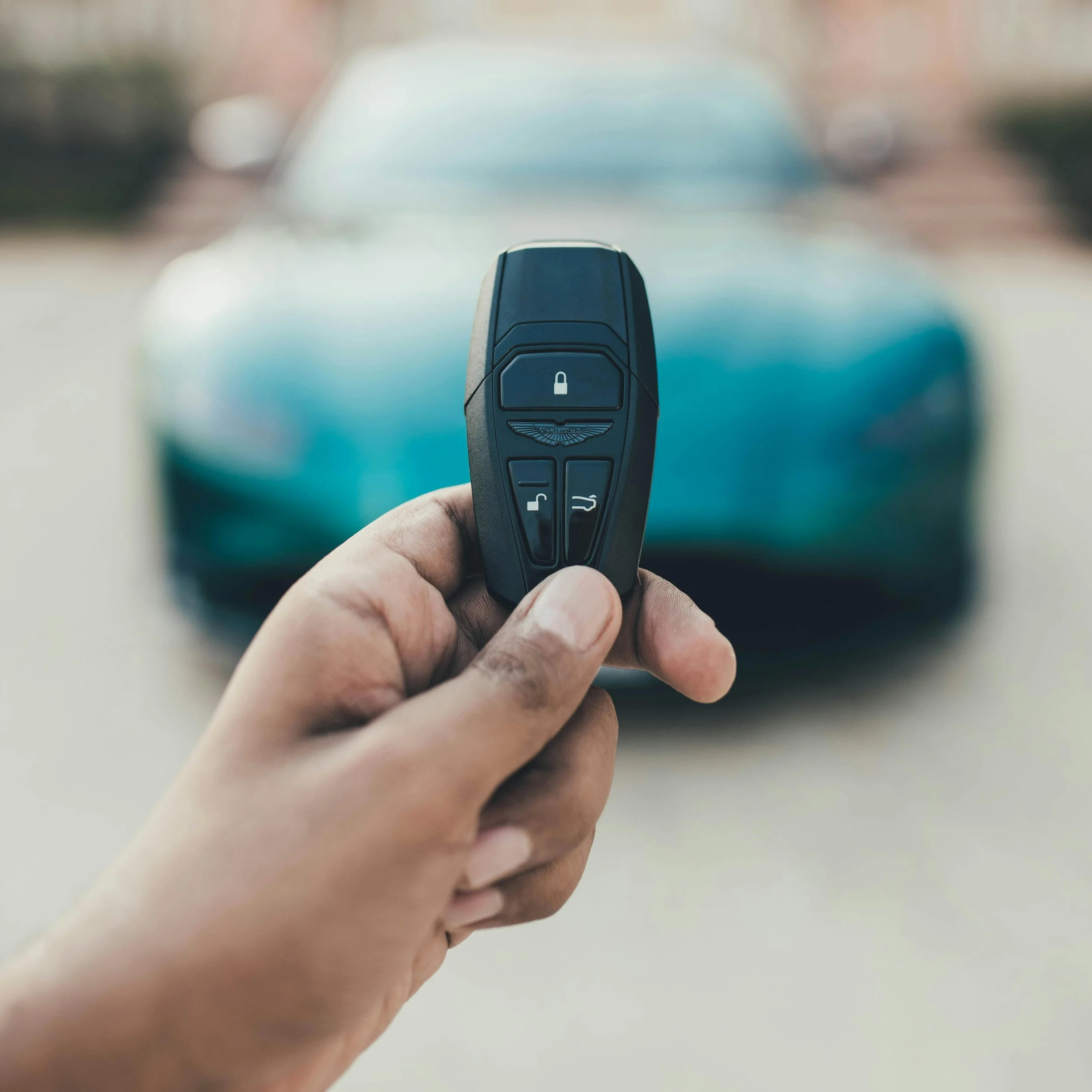 A close-up of a person's hand holding a black car key fob with a lock symbol on the button, with a blurred background.