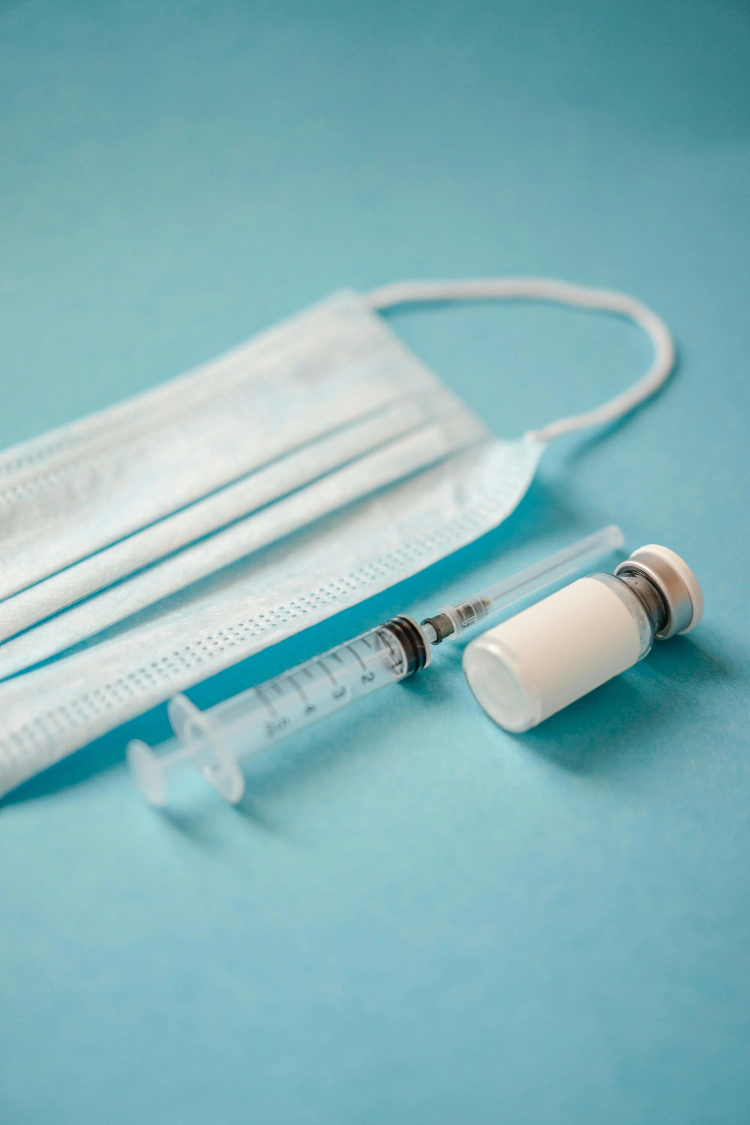 Close-up of a disposable face mask, syringe, and vaccine vial on a light blue background, related to medical or healthcare setting.