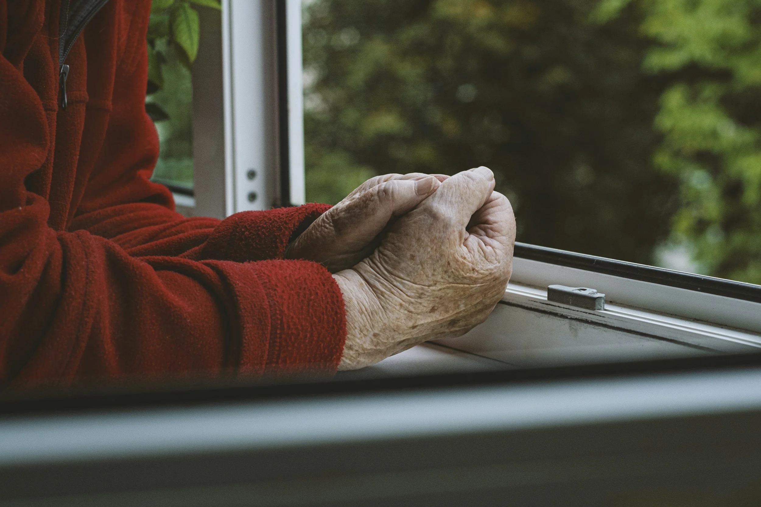 Close-up of elderly hands resting on a windowsill, with the background showing green blurred outdoor foliage, and part of a red fleece garment is visible.