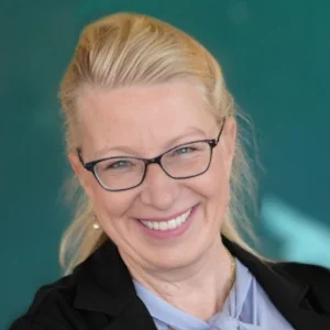 A smiling woman with blonde hair, glasses, and a black blazer, posing against a teal background.
