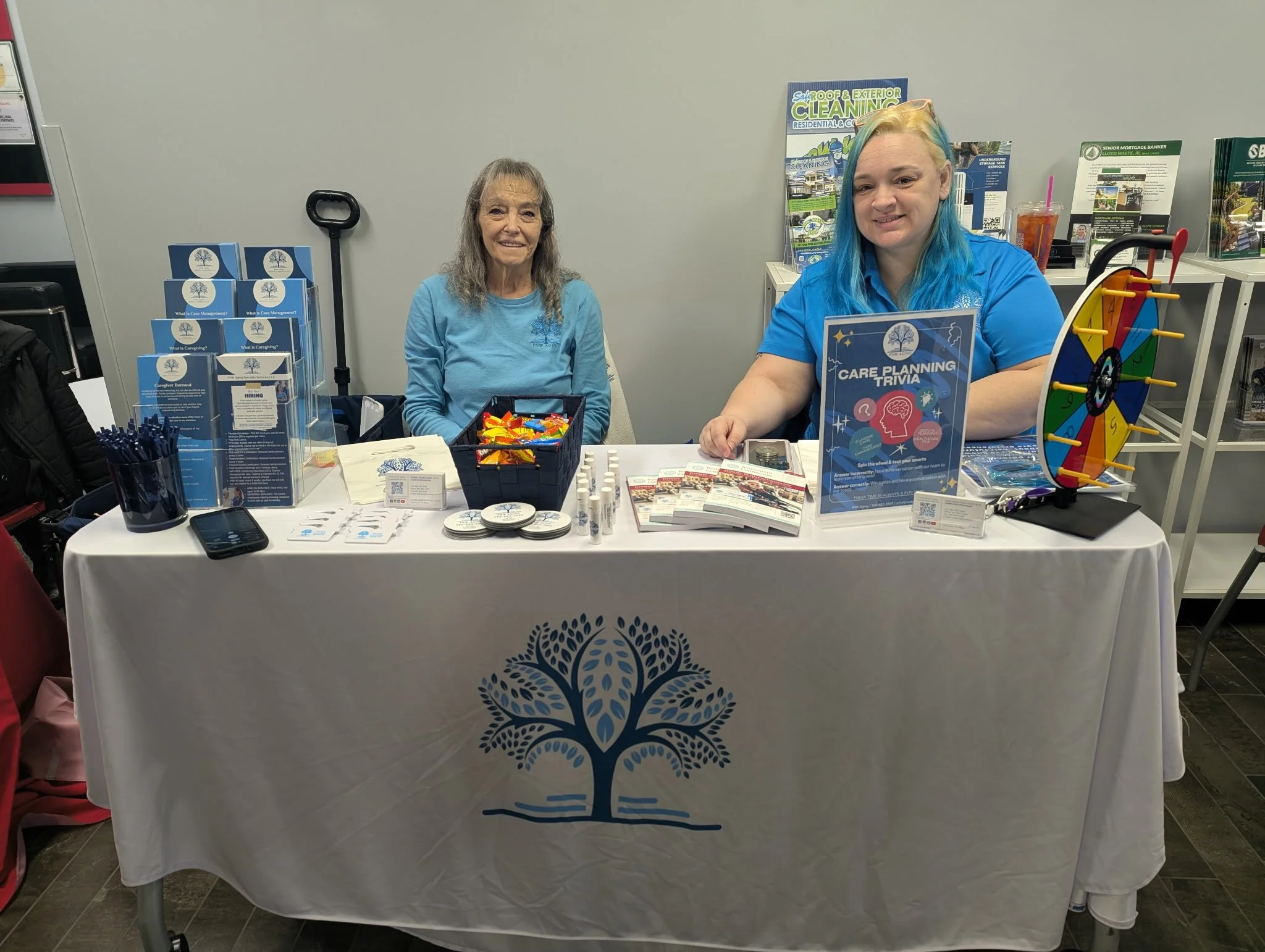 Two women sitting behind a table at a community event or fair, with promotional materials, a game wheel, and informational brochures.