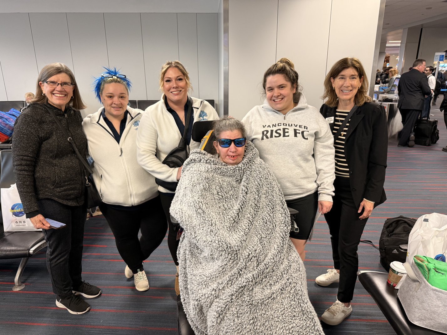 Group of six women and one person in a wheelchair posing together at an indoor airport for relocation support services. The individual in the wheelchair is covered with a blanket and wearing sunglasses.