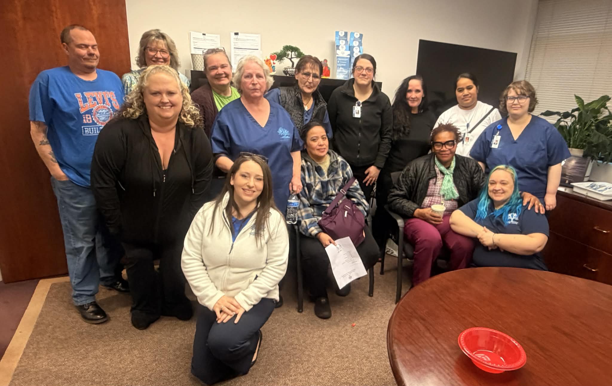 Group of diverse caregivers posing together in an office setting for training, with some seated and others standing, smiling at the camera.