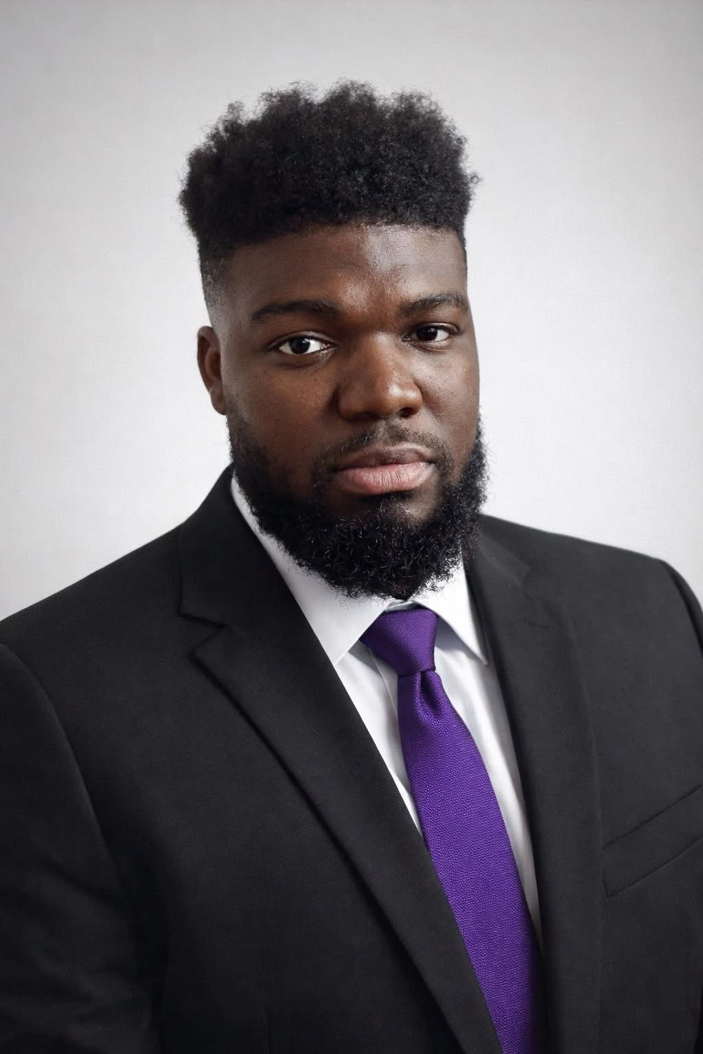 A young Black man in a business suit, white shirt, purple tie, looking at the camera with a serious expression, against a plain white background.