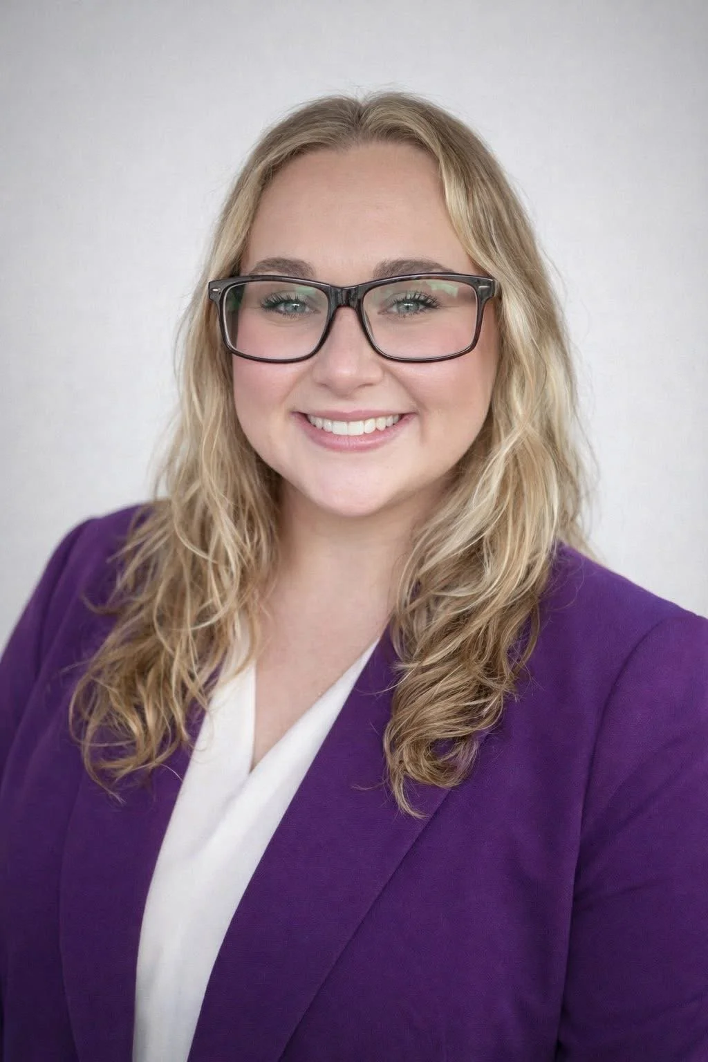 A woman with blonde, wavy hair wearing glasses, a purple blazer, and a white blouse, smiling against a plain background.