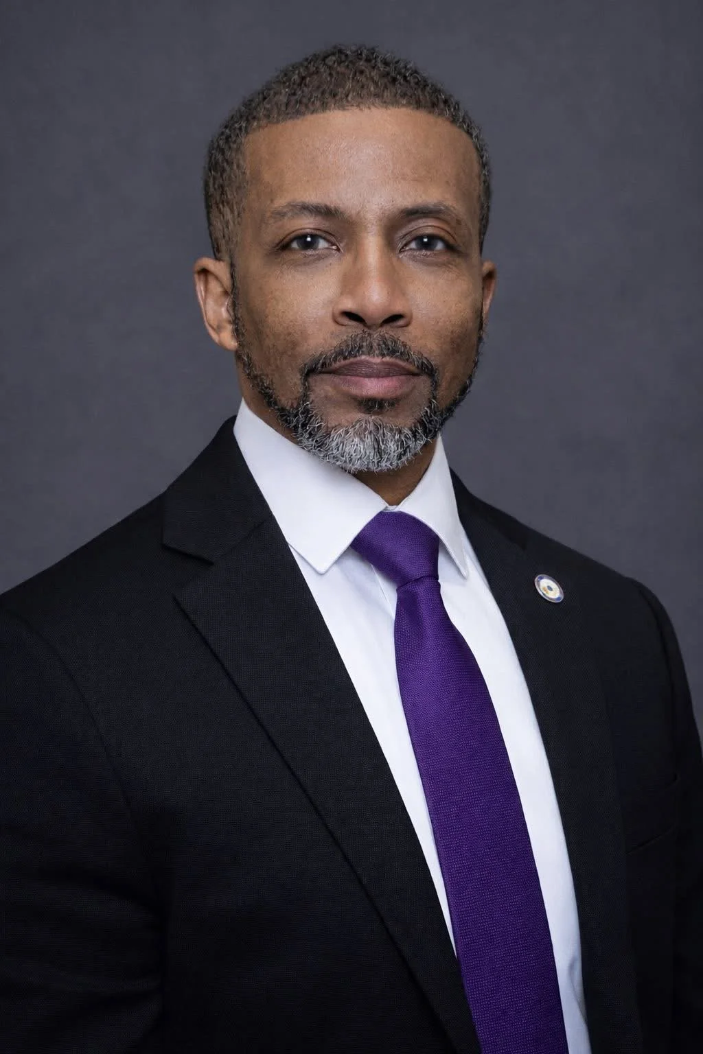 A professional portrait of a middle-aged African American man with short curly hair and a beard, wearing a black suit, white shirt, purple tie, and an American flag pin on his lapel, against a dark gray background.
