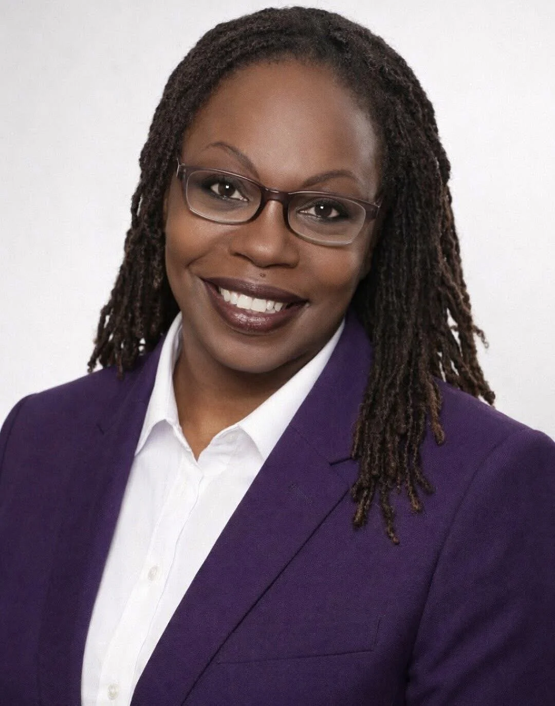 Portrait of an African American woman wearing glasses, a white shirt, and a purple blazer, smiling with white teeth against a plain white background.