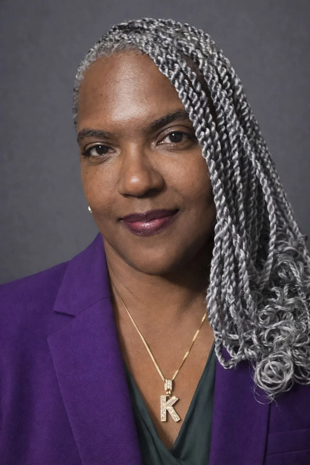 Portrait of a woman with silver box braids, wearing a purple blazer, gold necklace with a letter 'K', and a dark green top, against a gray background.