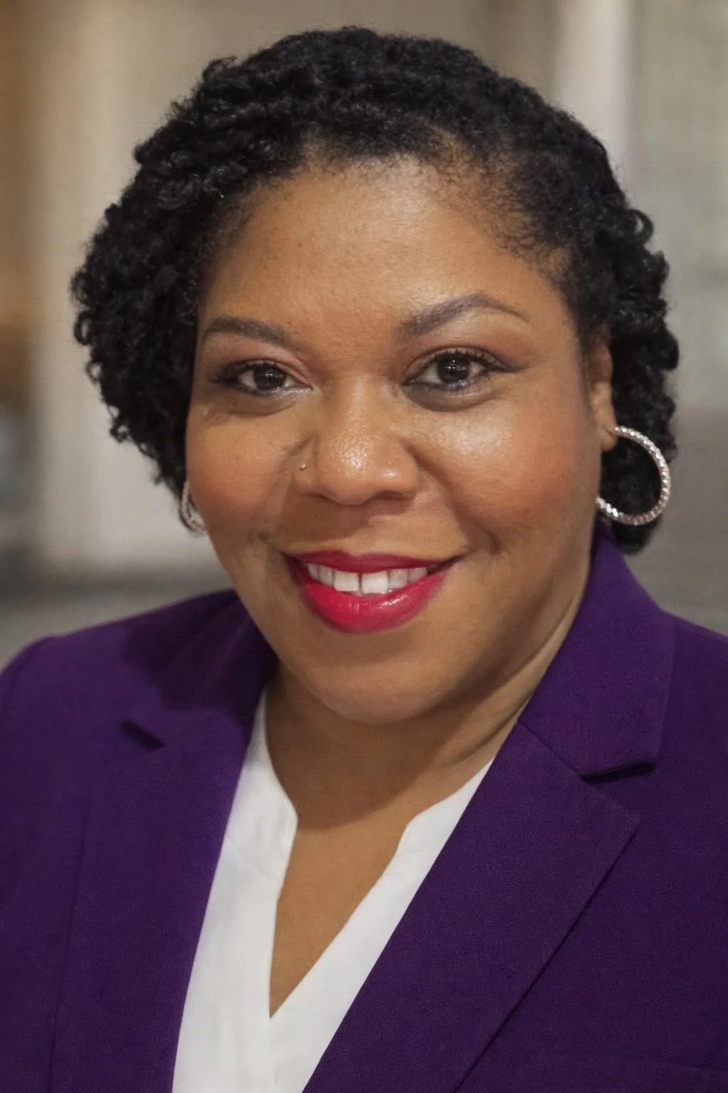 A woman with short curly black hair, wearing a purple blazer and hoop earrings, smiling at the camera.